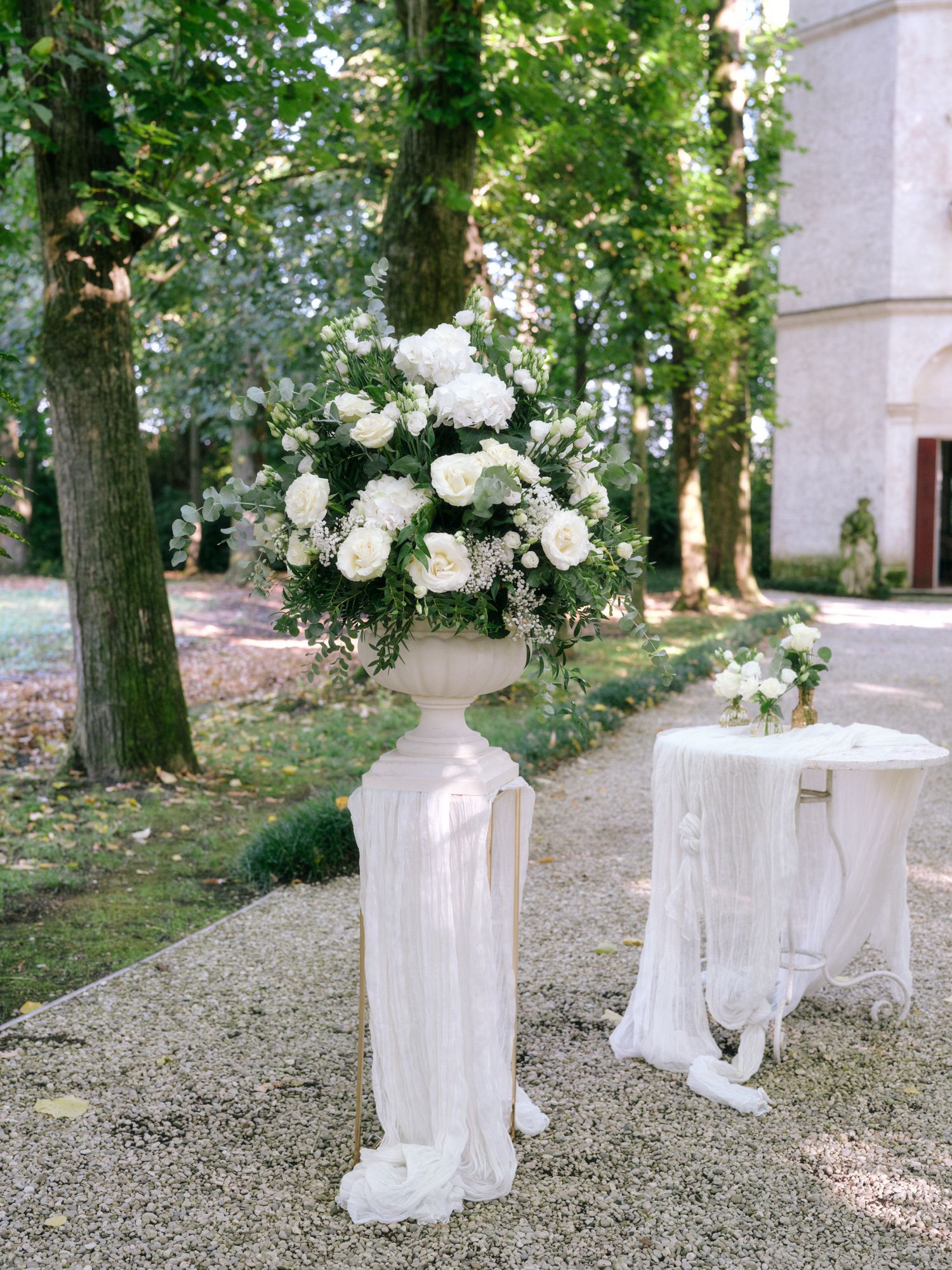 Elegant white floral arrangement on draped pedestal for outdoor garden wedding ceremony.