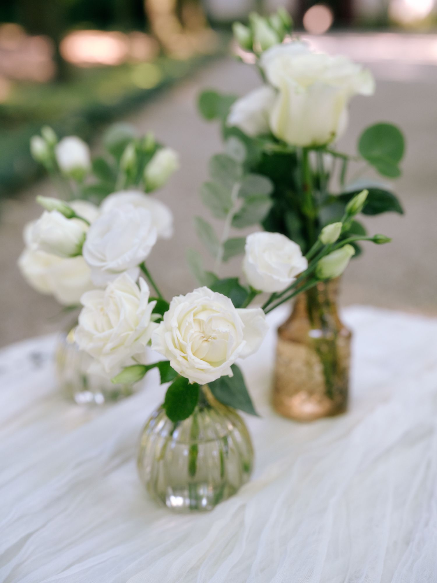 White roses and greenery arranged in clear and amber glass vases on a white textured surface.