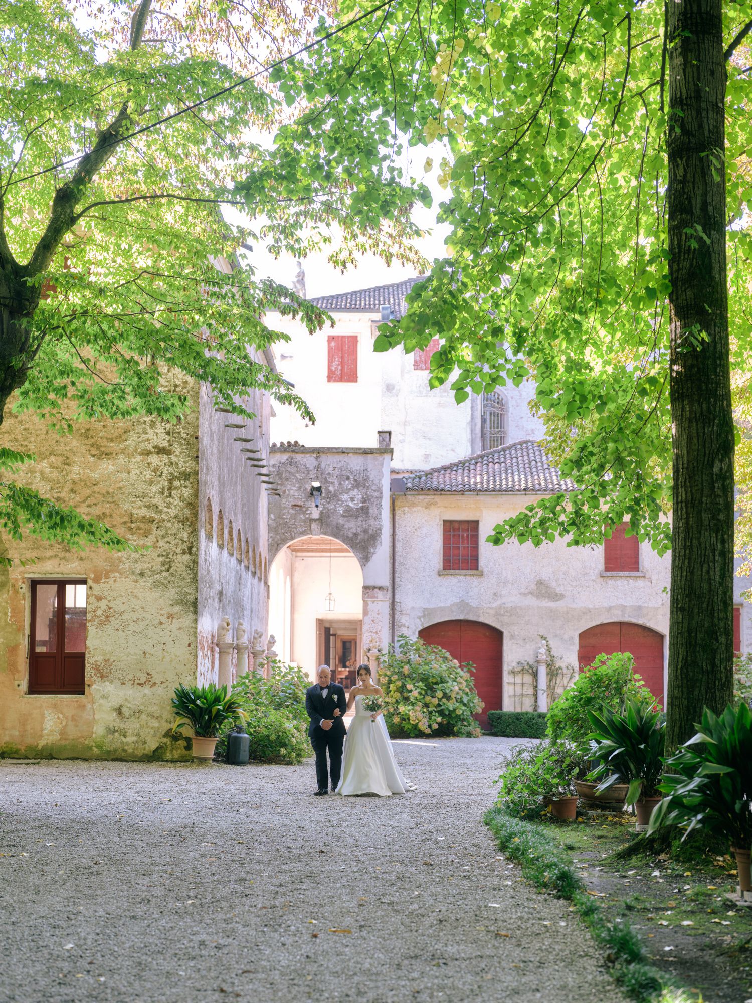 A couple walks along a gravel path through a picturesque Italian villa courtyard framed by green trees and white walls.
