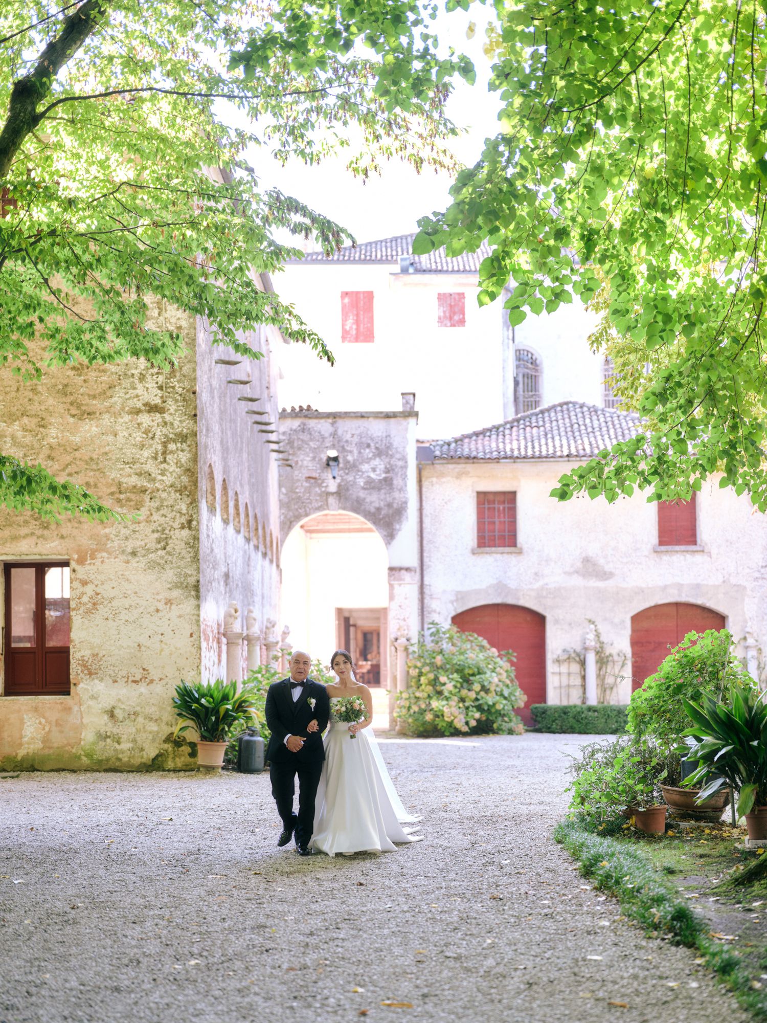 A couple stands in a romantic stone courtyard surrounded by lush green trees and historic architecture in Italy.