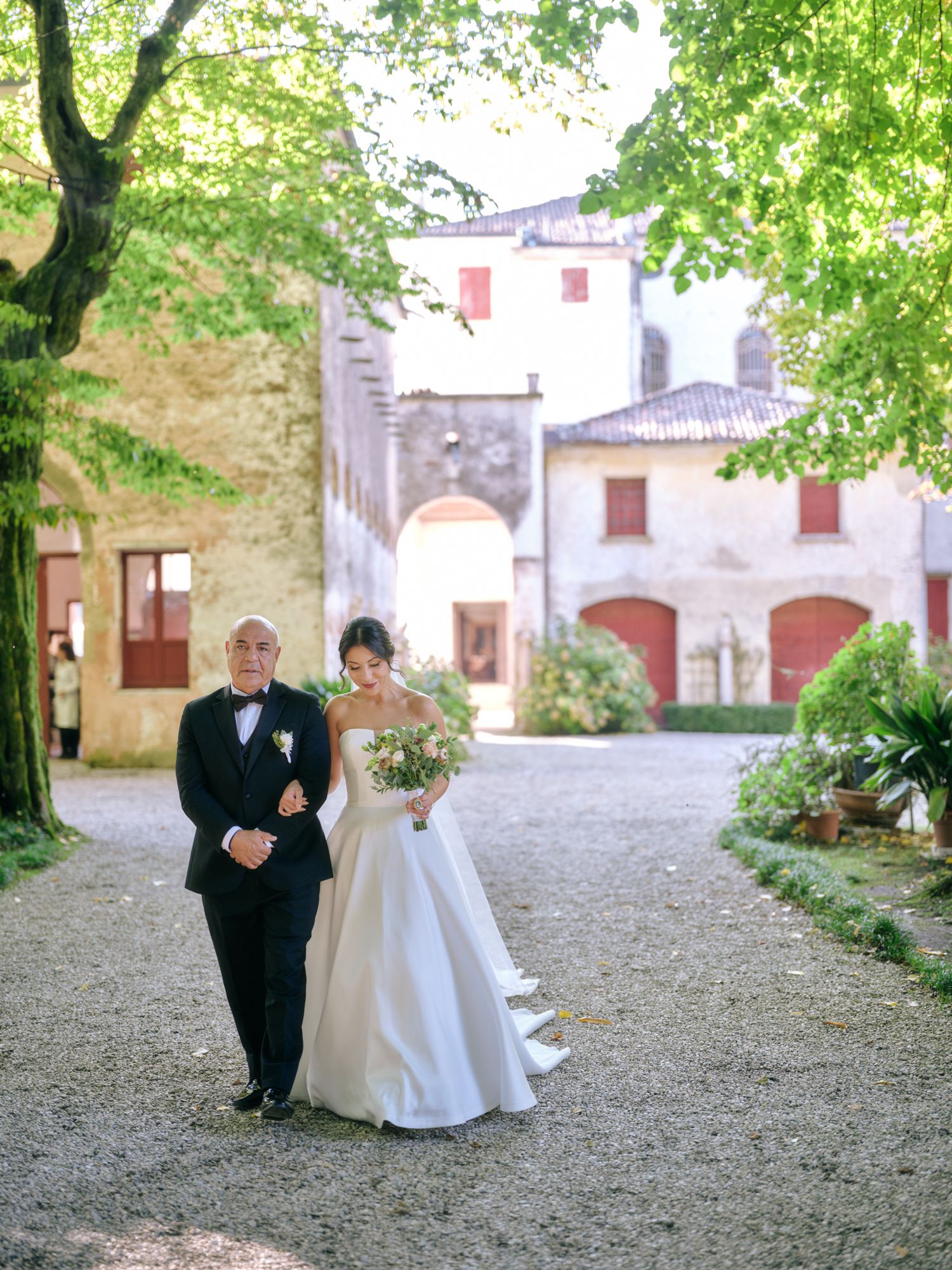 A couple poses for wedding photos in a rustic courtyard setting surrounded by green trees and historic buildings.