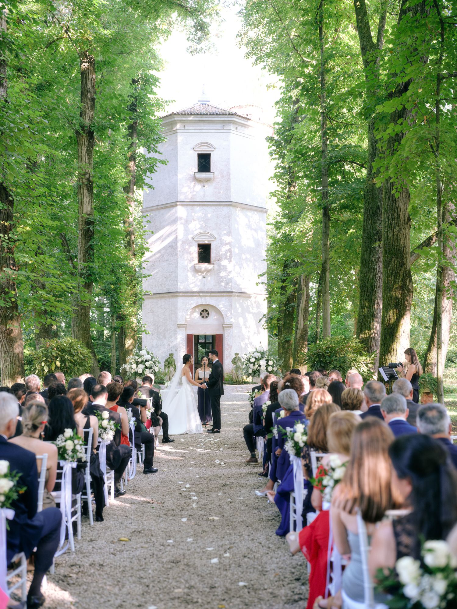 An outdoor wedding ceremony takes place in front of a white chapel tower surrounded by tall green trees.