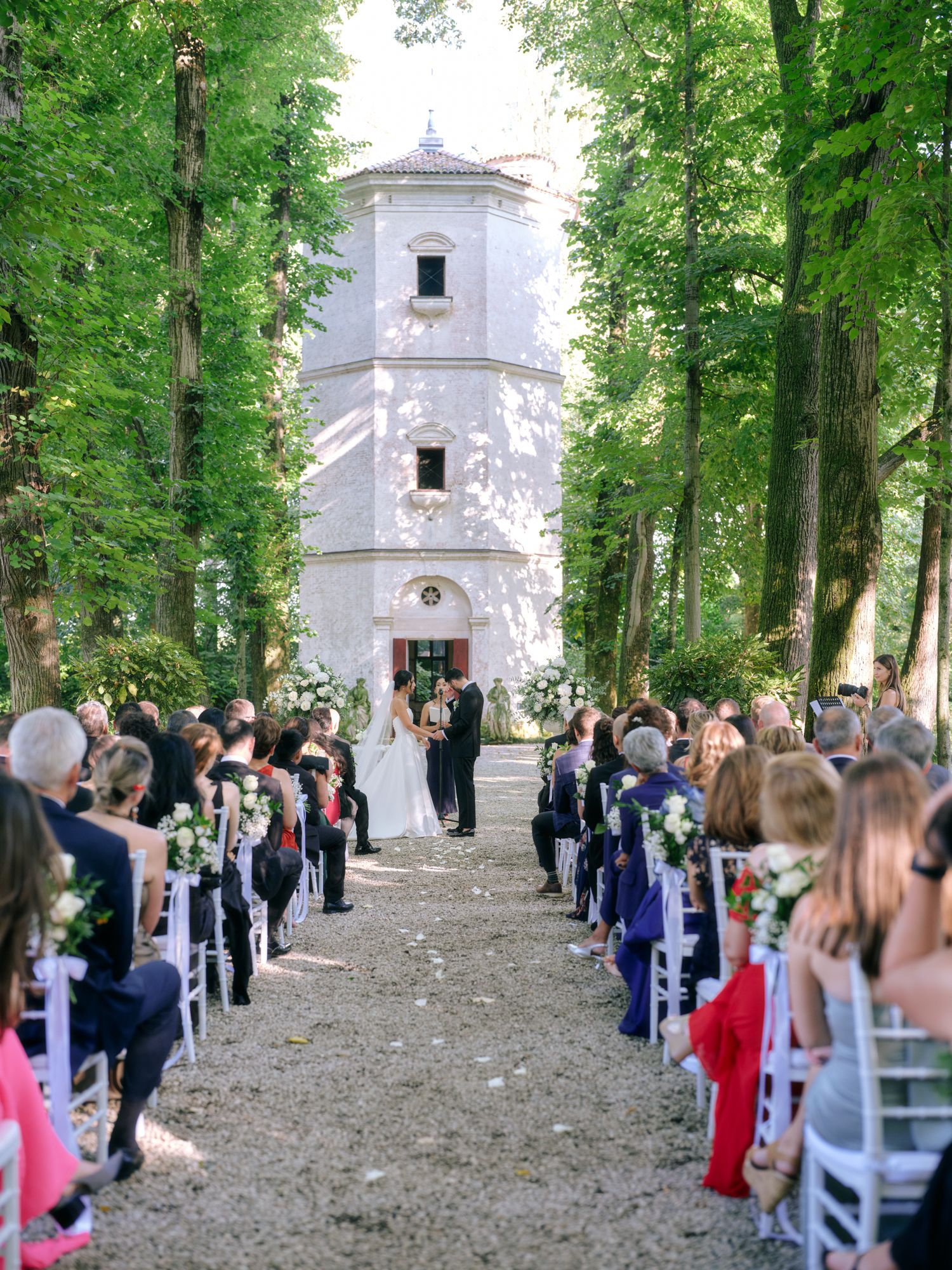 Wedding guests gather along a tree-lined path leading to a white chapel for an outdoor ceremony.