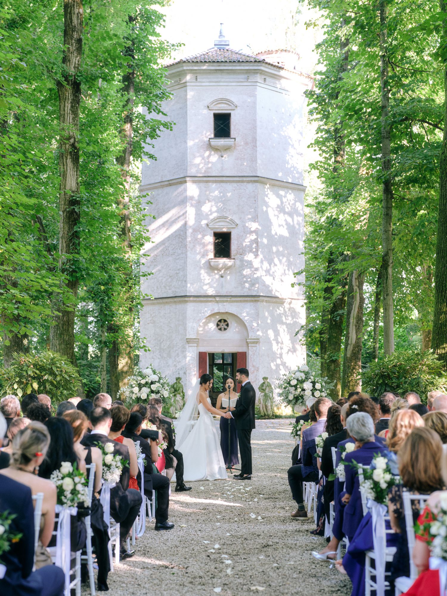 A wedding ceremony taking place outdoors near a white stone chapel surrounded by tall green trees.