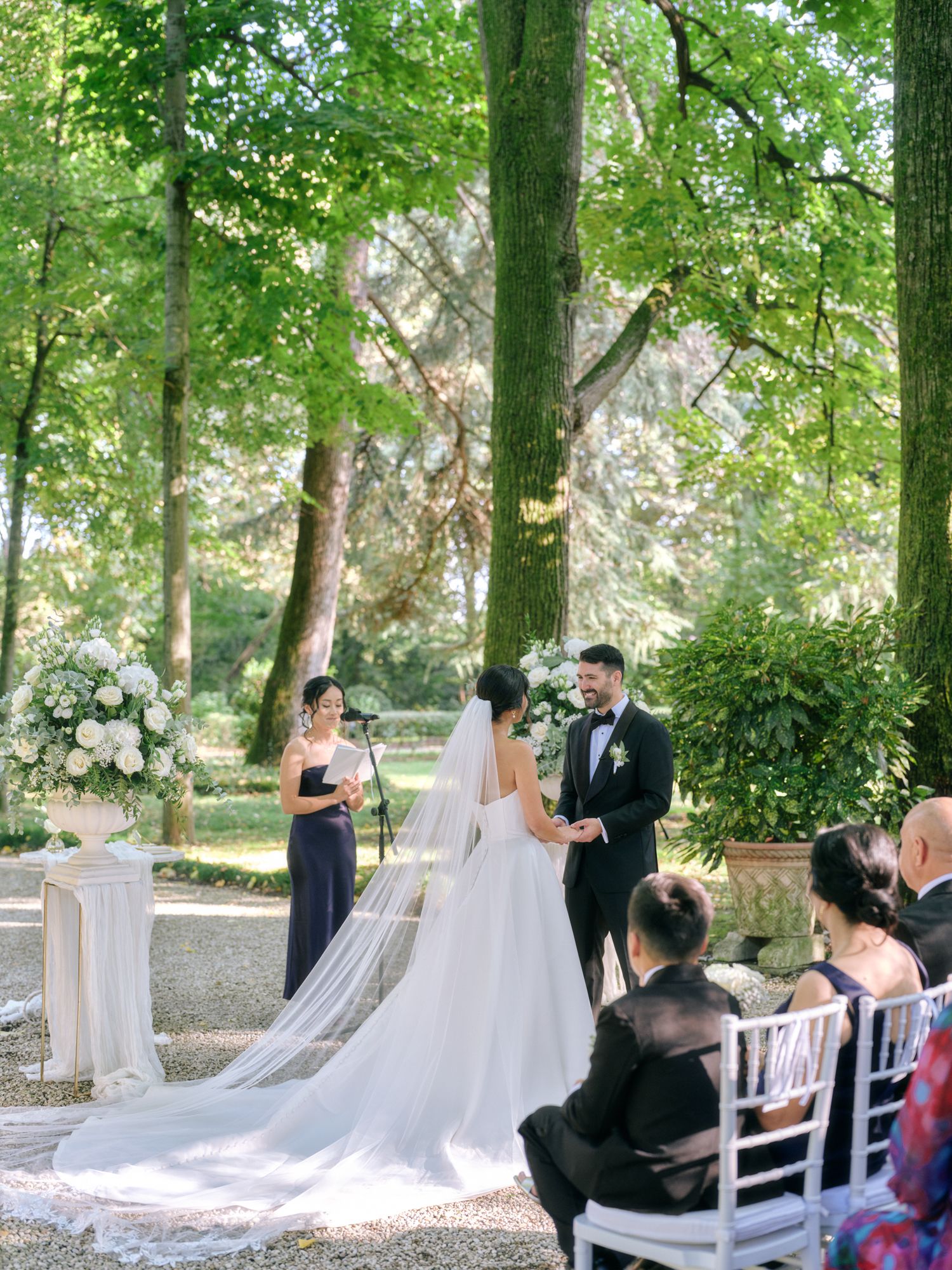 A wedding ceremony takes place in a serene outdoor garden setting with flowing white veils and natural greenery.
