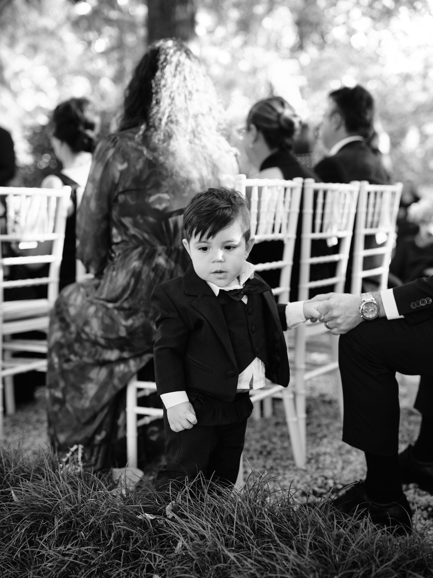 Black and white photo of a young boy in formal attire looking unamused at an outdoor wedding with white chairs.