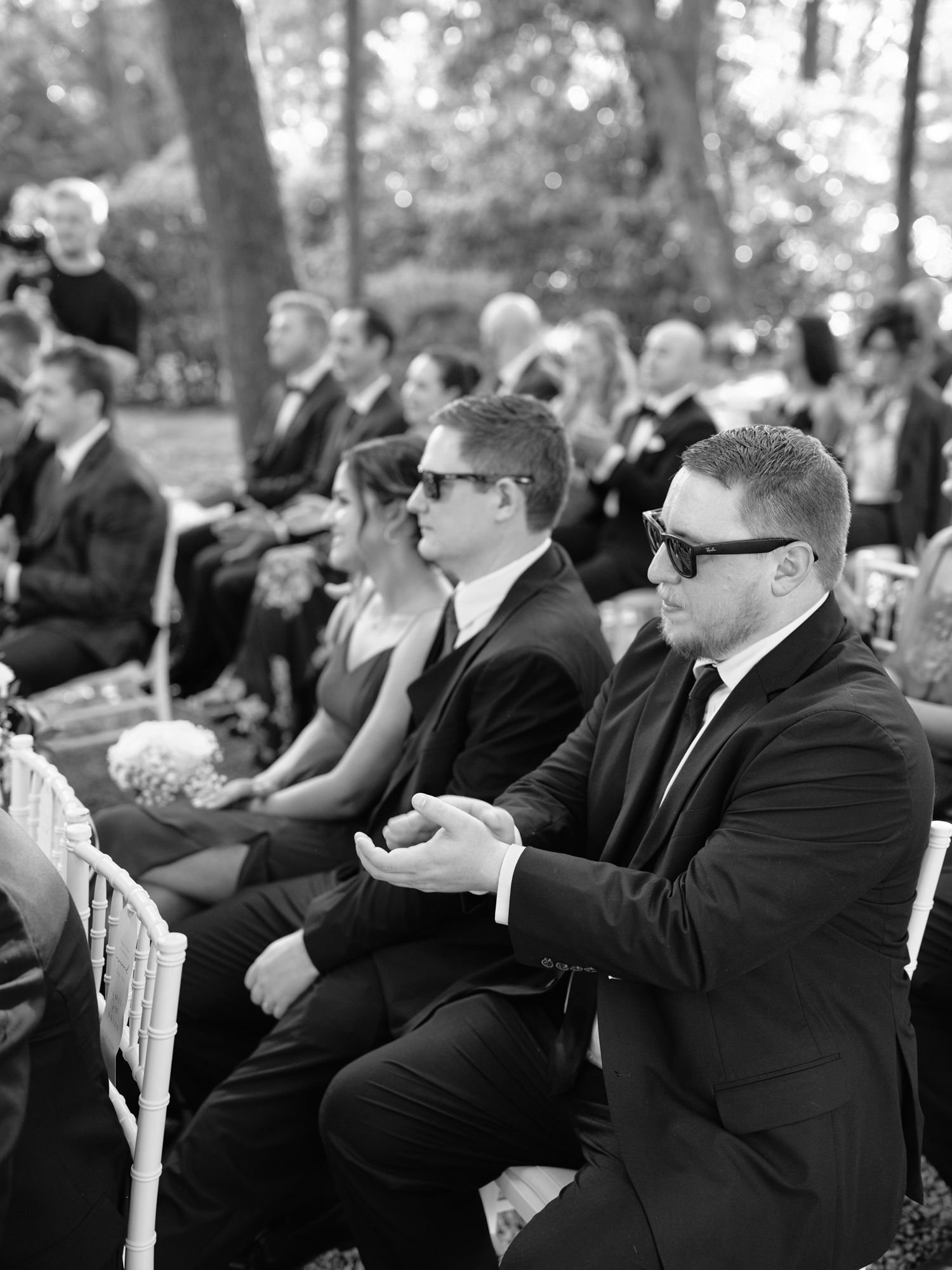 Wedding guests in formal attire seated on white chairs during an outdoor ceremony.