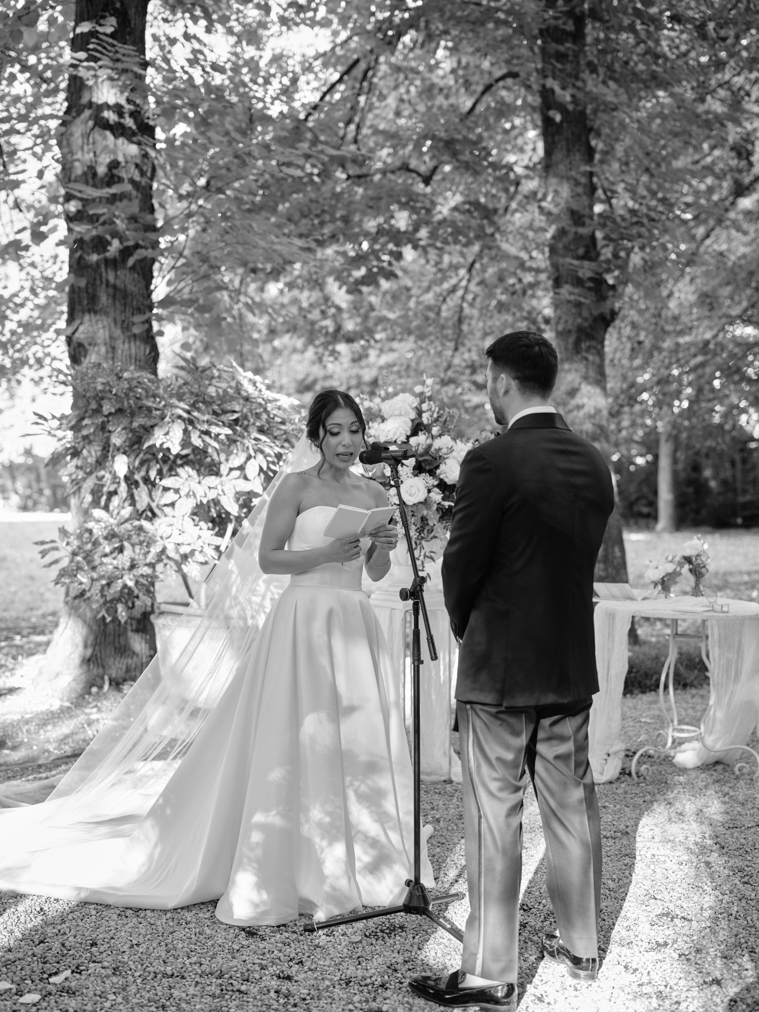 A couple in wedding attire poses together in a romantic outdoor garden setting with sunlit trees in the background.