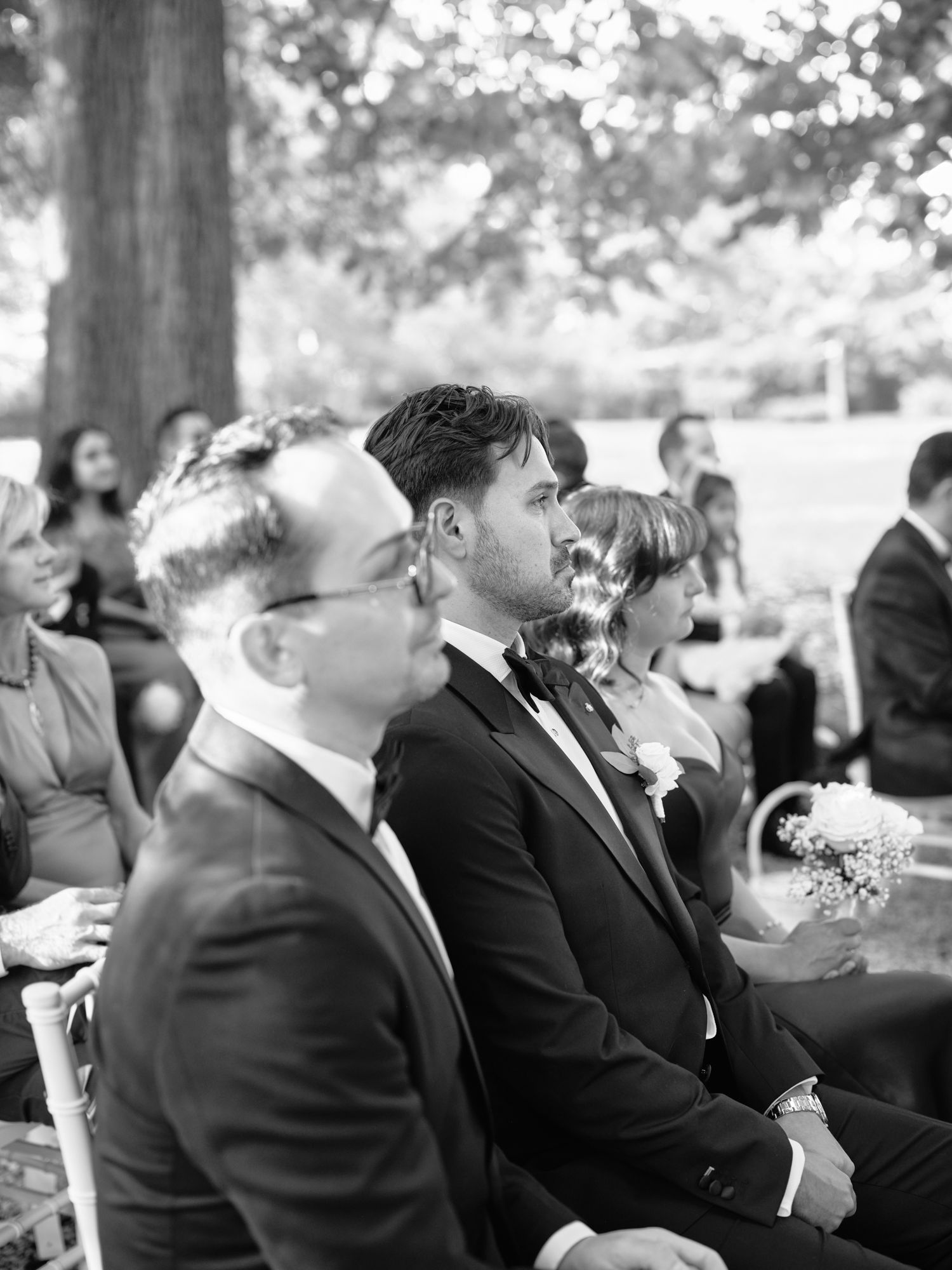 Black and white photo showing groomsmen in dark suits seated outdoors at a wedding ceremony.
