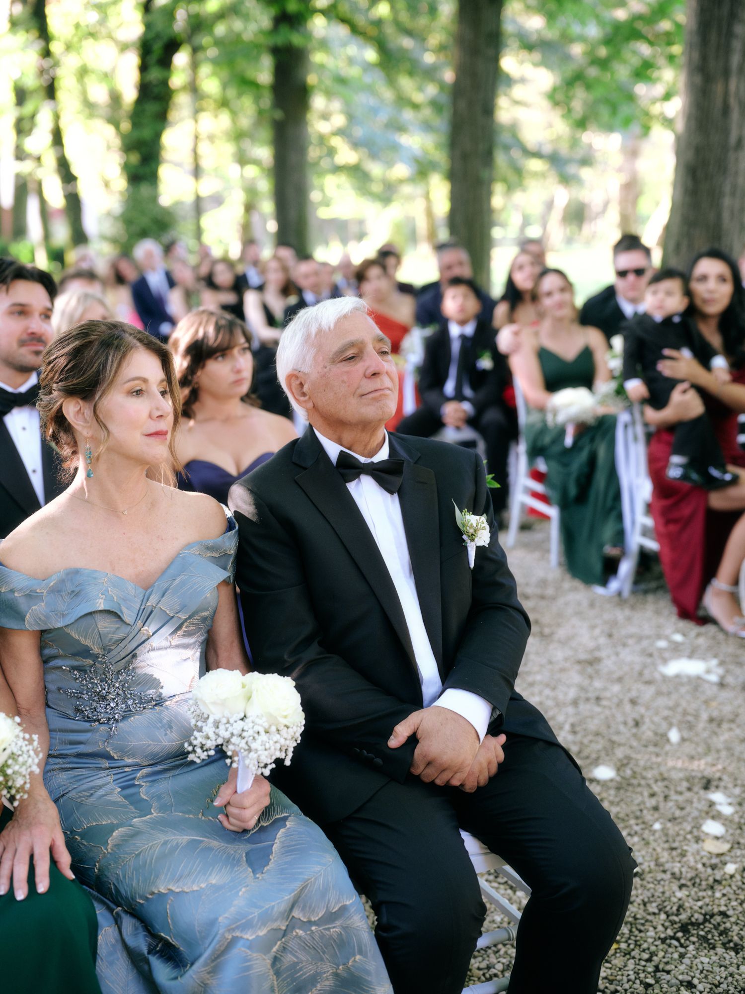 Wedding guests seated outdoors in formal attire watch a ceremony under a canopy of trees in a forest setting.