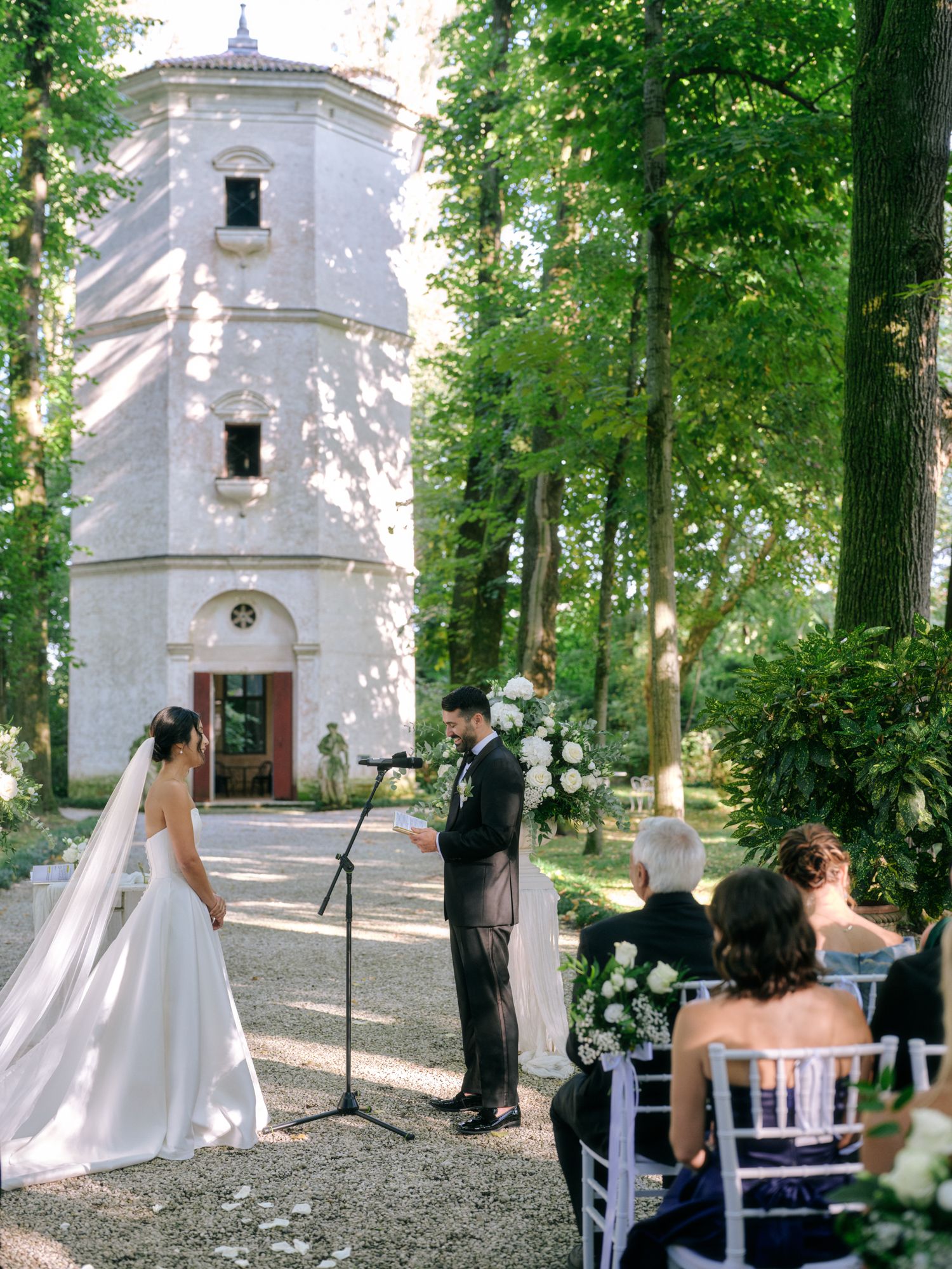 An outdoor wedding ceremony takes place in a wooded area with a white chapel in the background.
