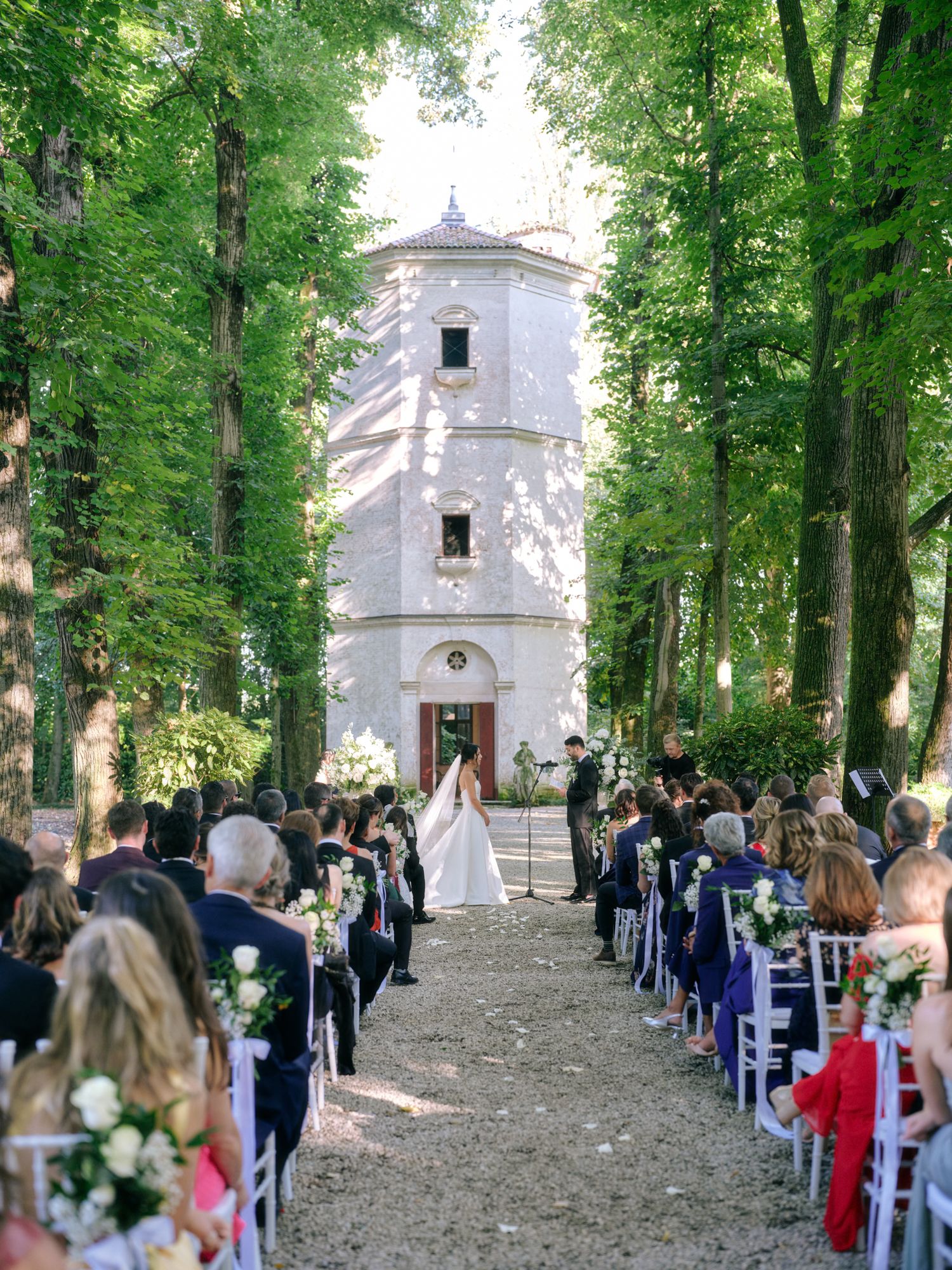 Guests gather along a tree-lined path leading to a white stone church for an outdoor ceremony in a wooded setting.