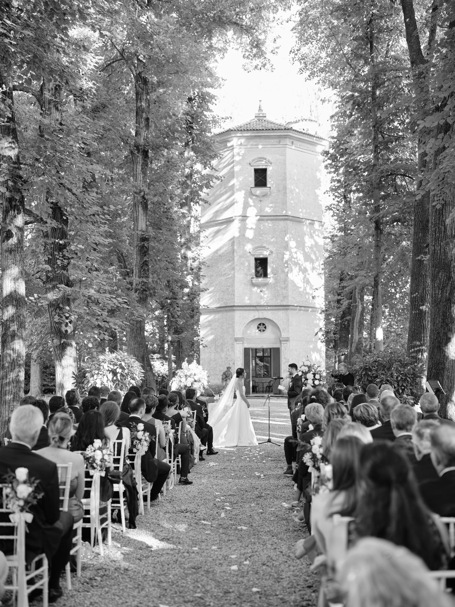 A black and white photograph shows wedding guests seated in rows among tall trees near a historic chapel.