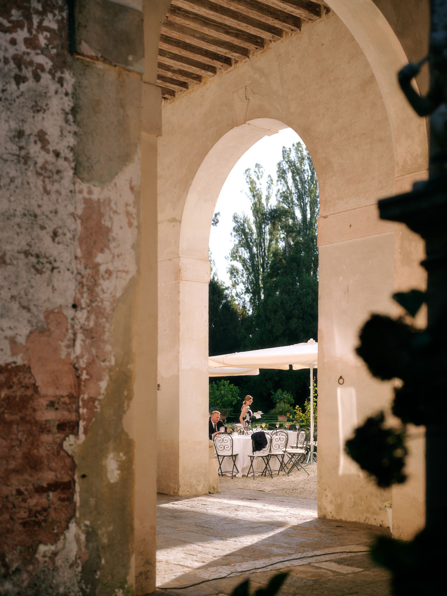 View through an arched stone doorway showing outdoor seating area with tables and chairs beneath tall trees.