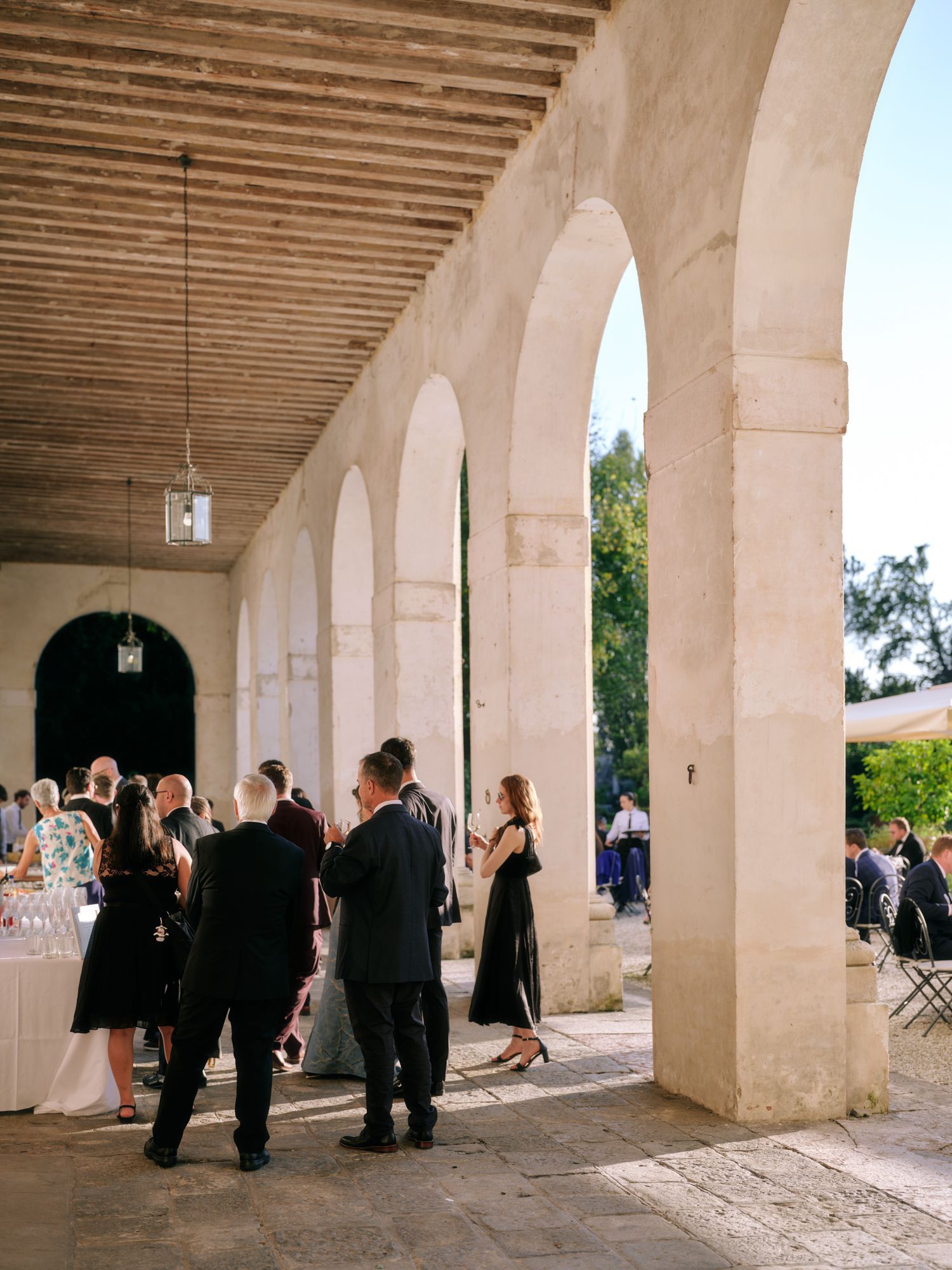 People gather under a covered colonnade with arched openings and wooden beamed ceiling at an outdoor venue.