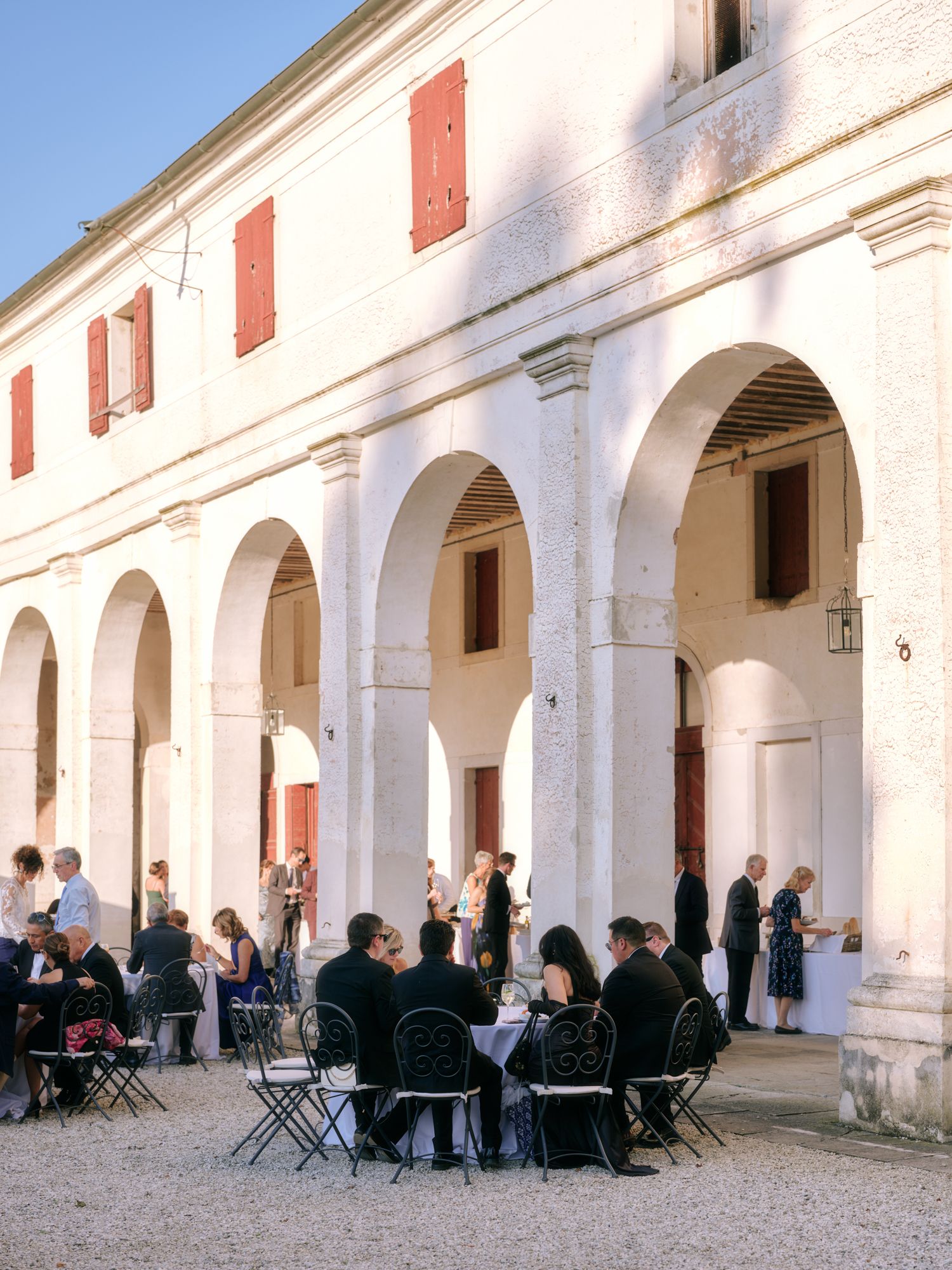 People gather in a white arched colonnade courtyard with red shuttered windows during a social event.