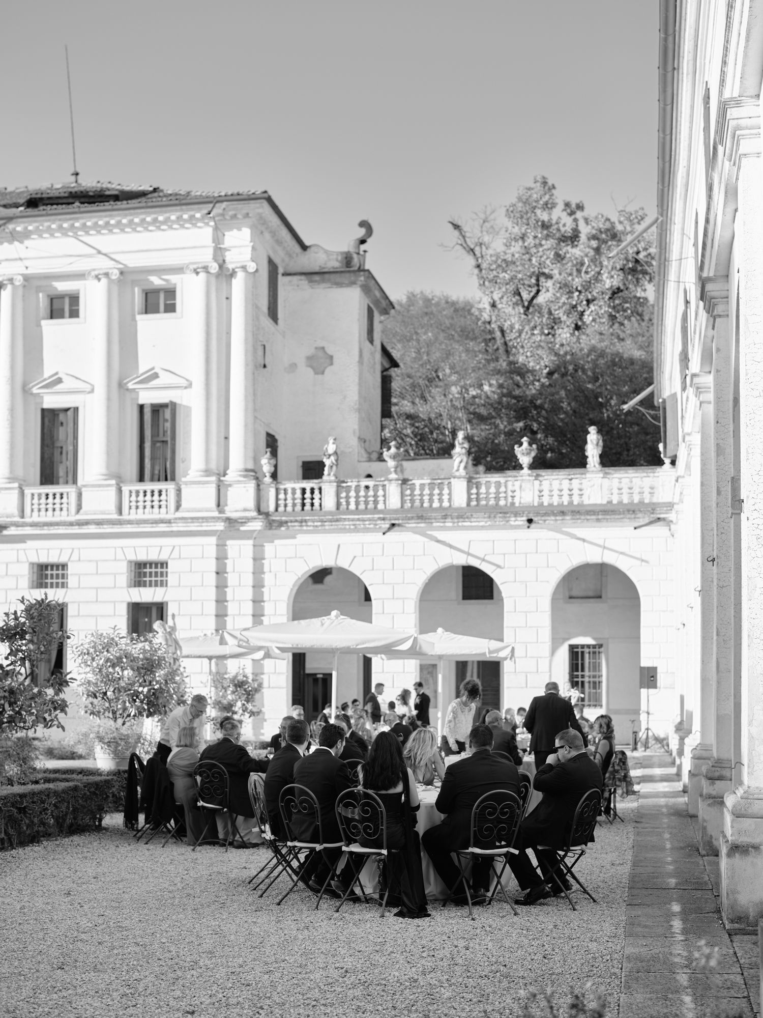 A group of people seated at round tables on a patio with white columns and arches in a Mediterranean setting.