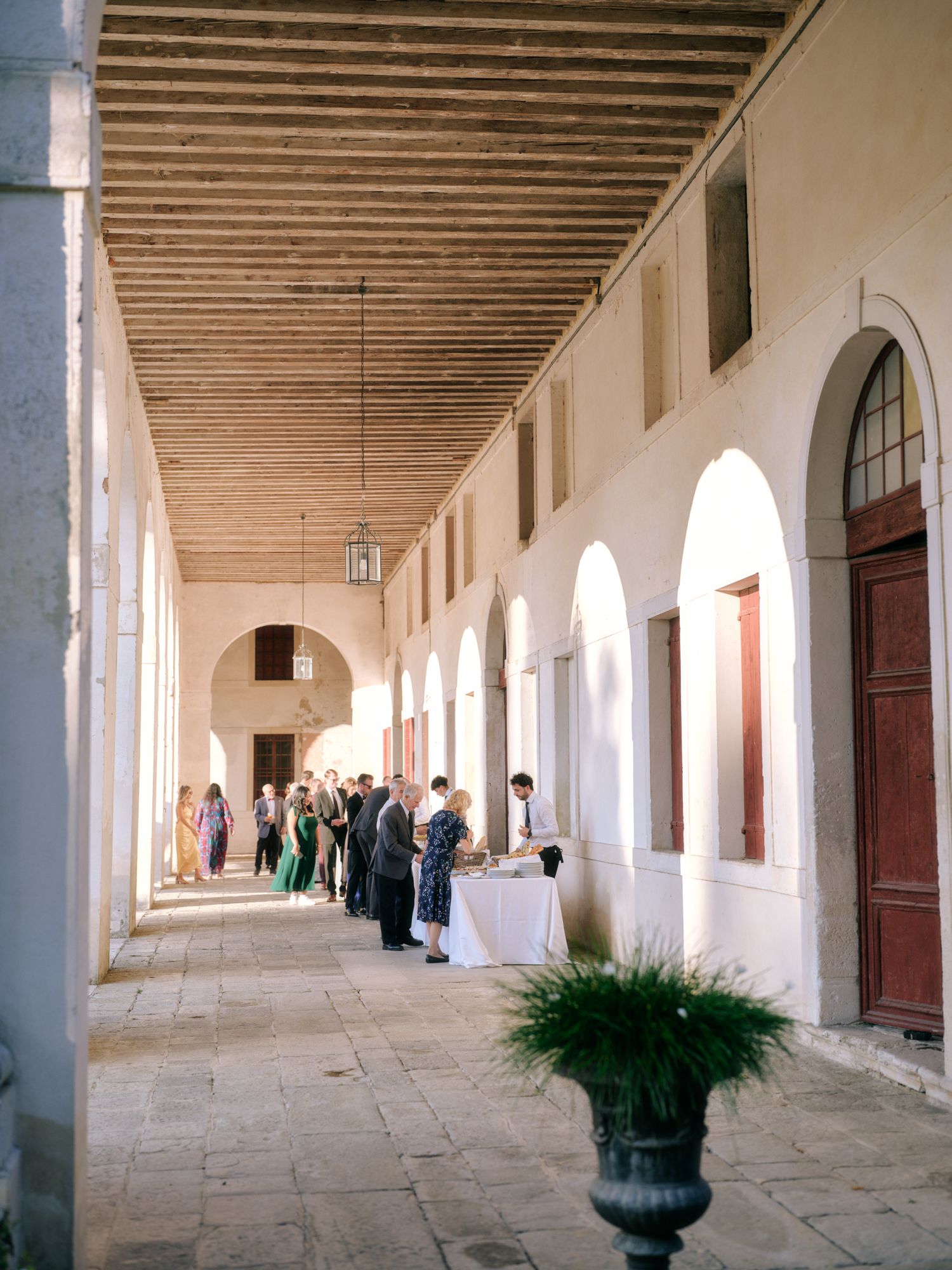 People gather in an elegant arched colonnade hallway with white walls and wooden beamed ceiling.