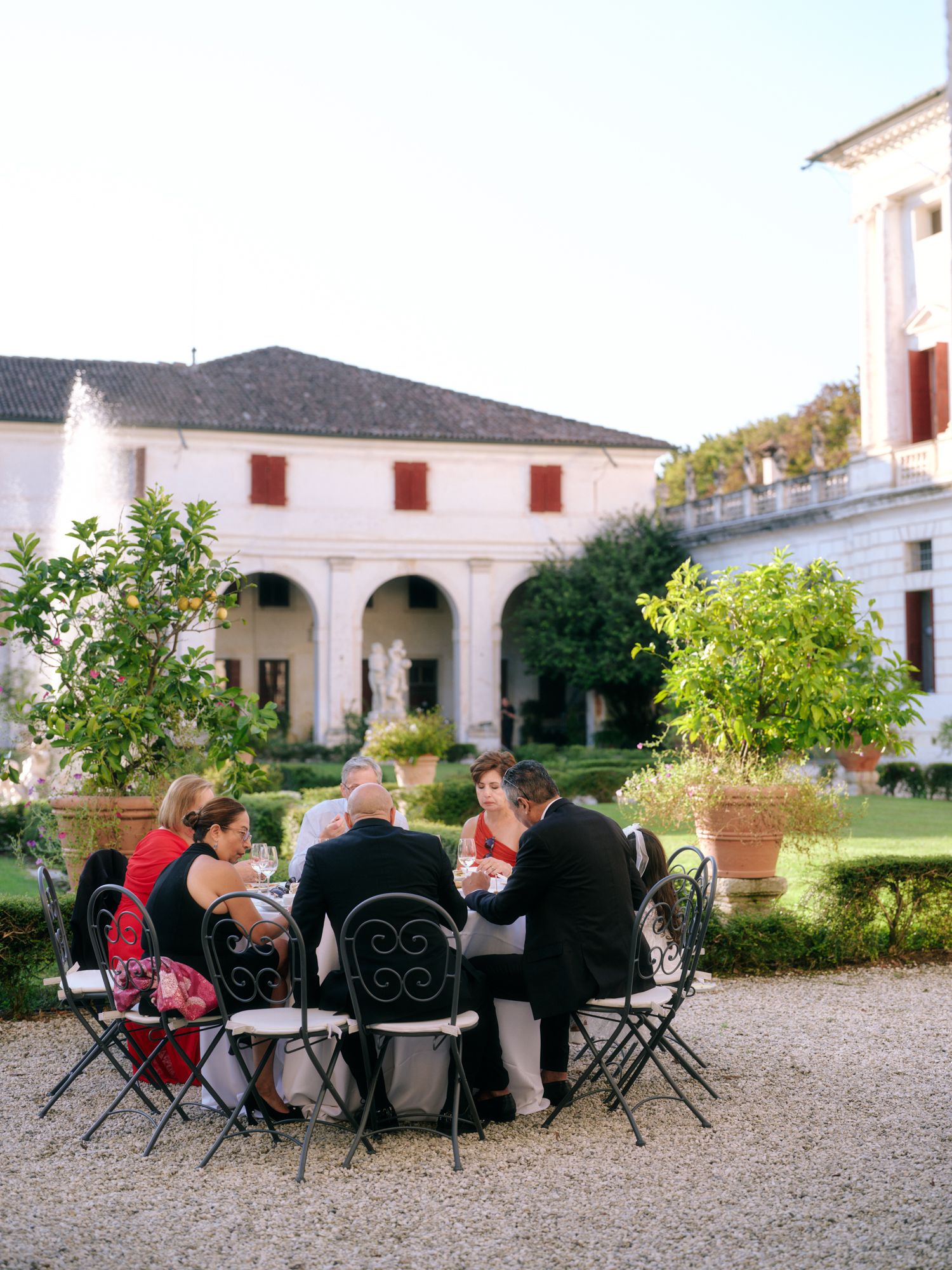 Groups sit in folding chairs on a gravel courtyard surrounded by white colonial architecture with red accents.