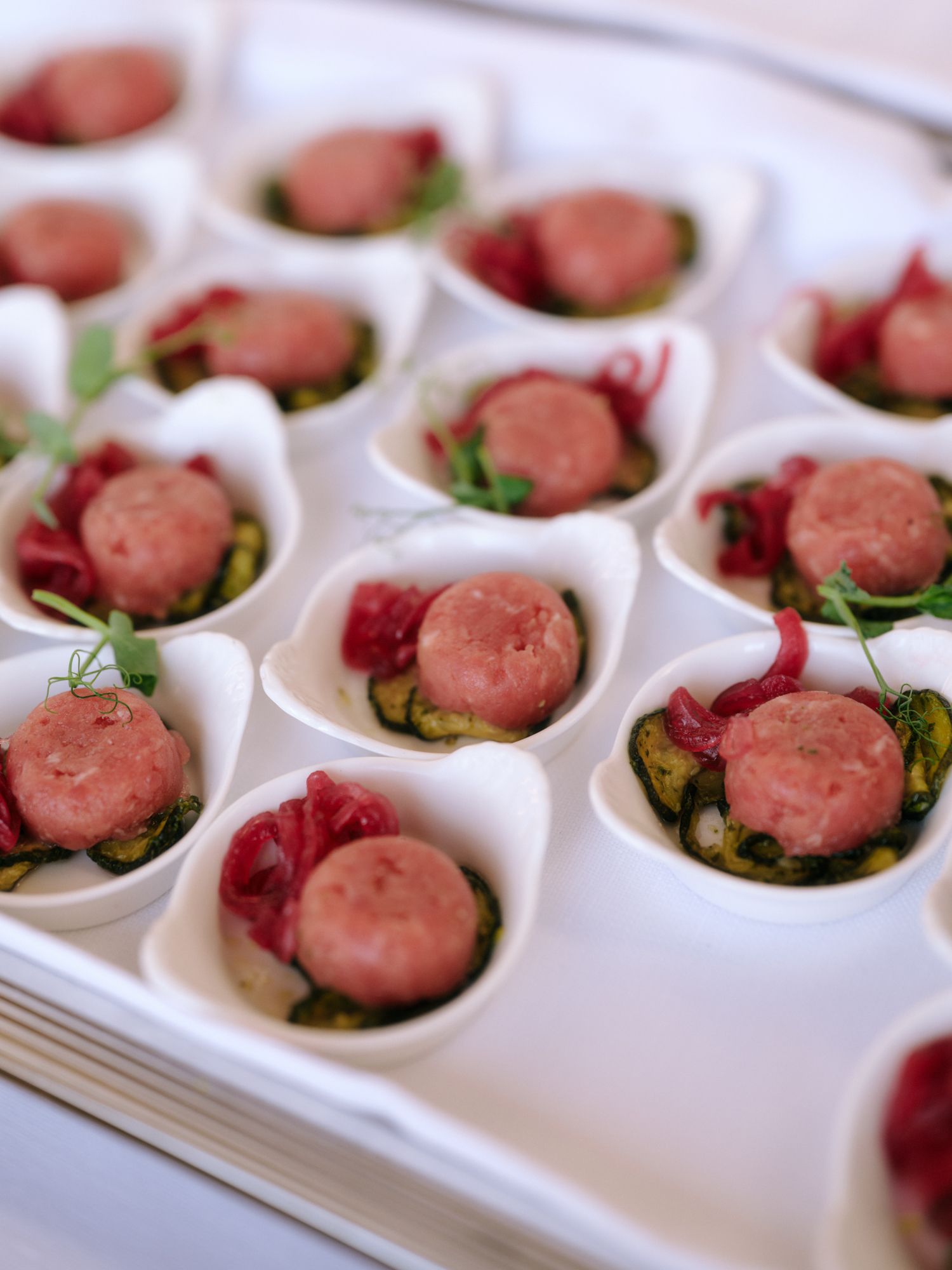 Small pink sausages served in individual white porcelain spoons with garnishes and red accents for elegant appetizers.