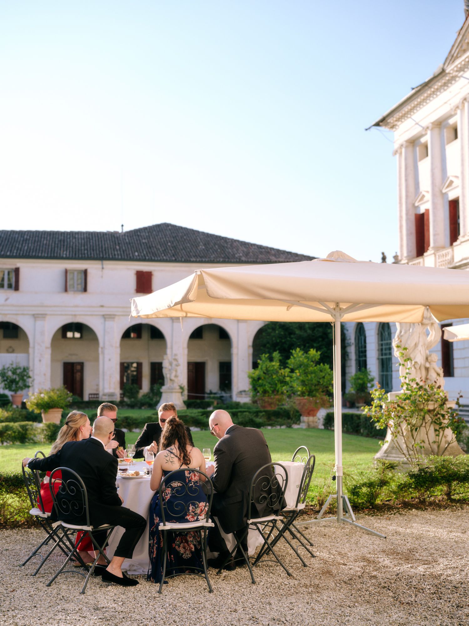 Group dining outdoors under a white umbrella in an elegant courtyard with arched columns and Mediterranean architecture.