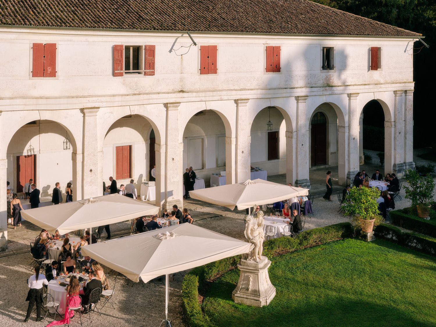 People gather under white umbrellas at an elegant Italian villa with arched colonnade and manicured lawn.