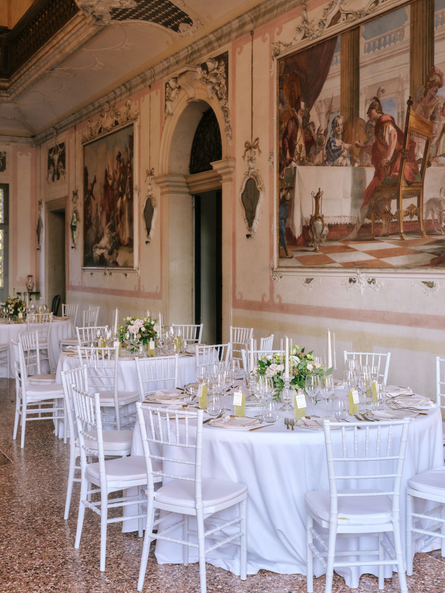 Elegant wedding reception setup with white Chiavari chairs and tables in a historic Italian villa with frescoed walls.
