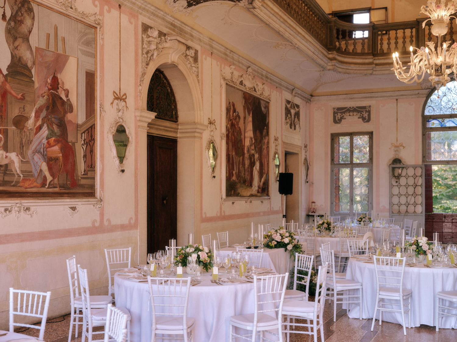 Elegant wedding reception setup in a grand ballroom with crystal chandeliers, white chairs and ornate wall decorations.