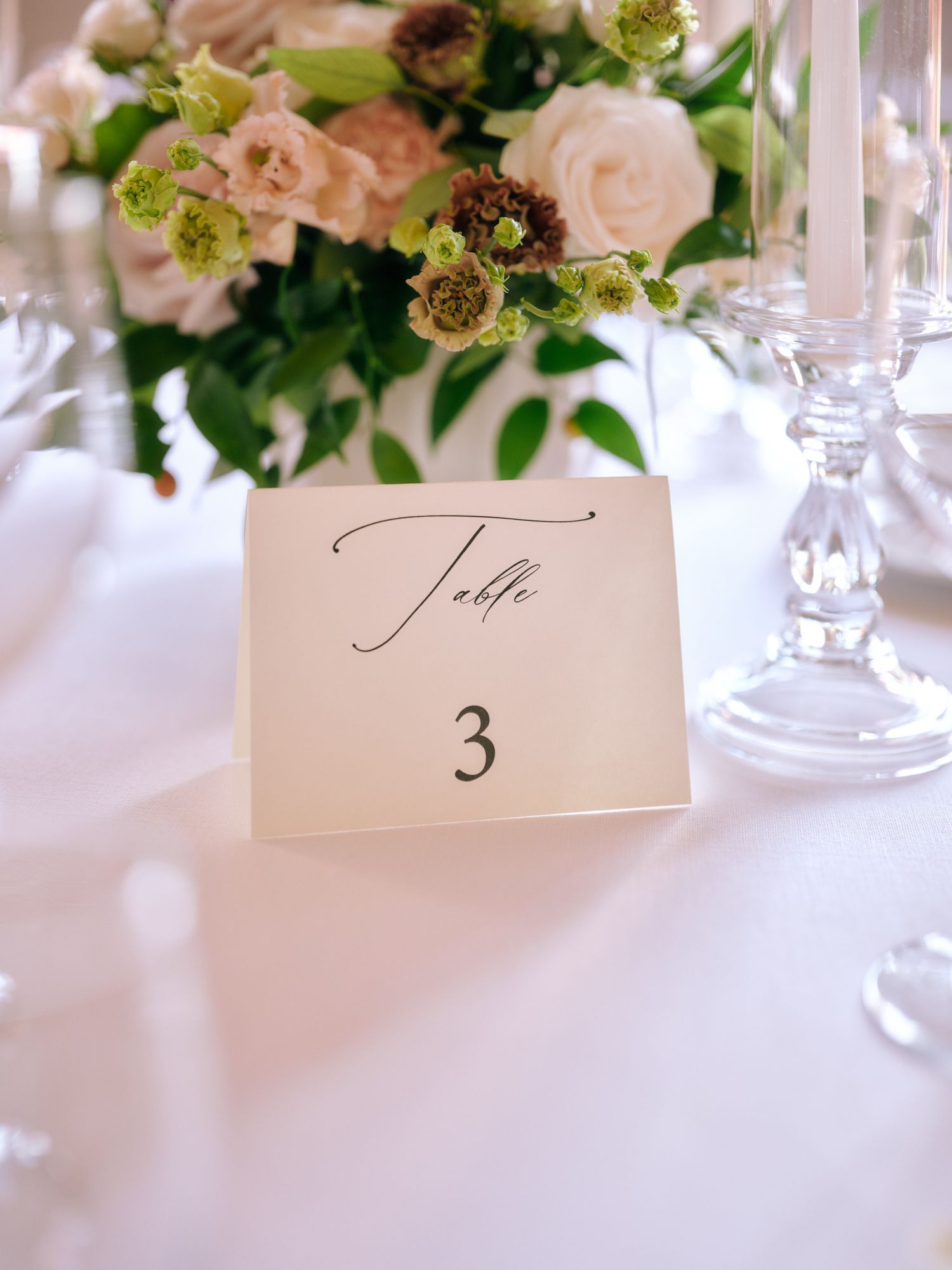 Elegant wedding table number 3 card displayed with pink roses and crystal glassware on white tablecloth.