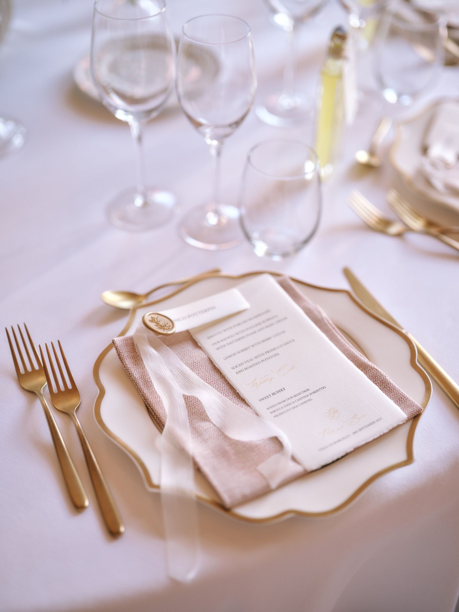Elegant place setting with gold flatware, white plates, wine glasses and menu card on crisp white tablecloth.