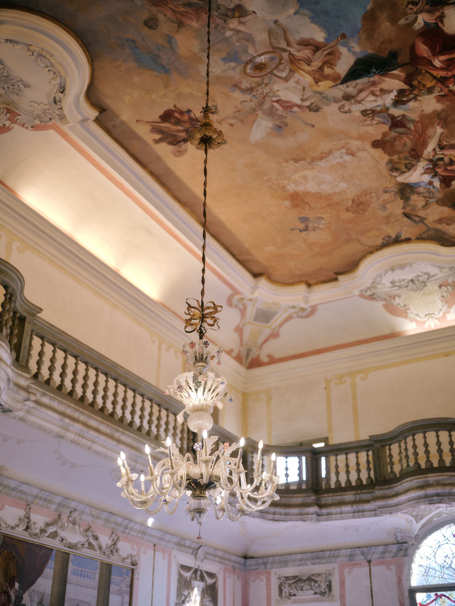 Ornate baroque ceiling with fresco paintings and crystal chandeliers in an elegant palace interior with white balustrades.