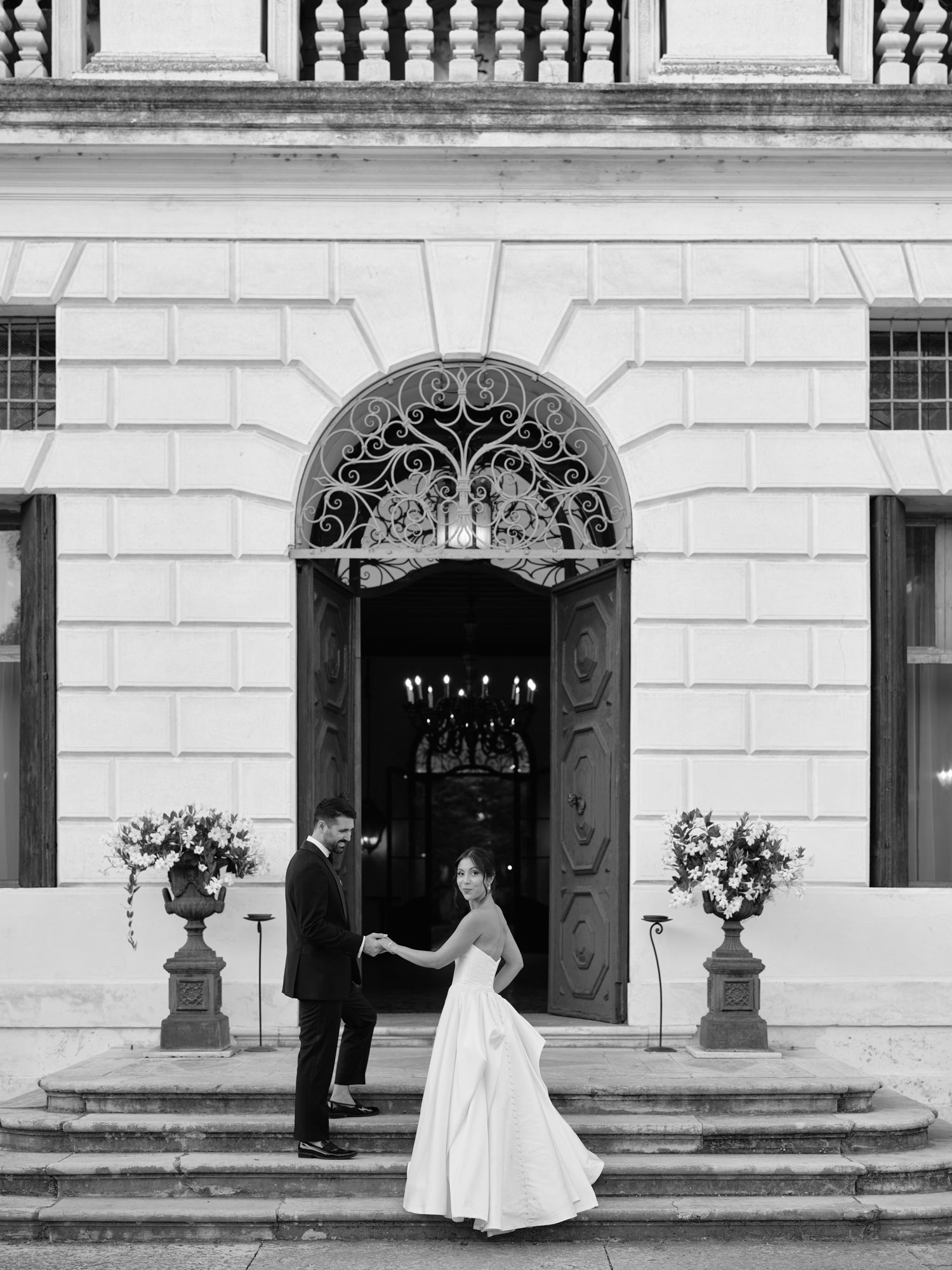 A wedding couple and guests stand outside an elegant white building with ornate doorways and classical architecture.
