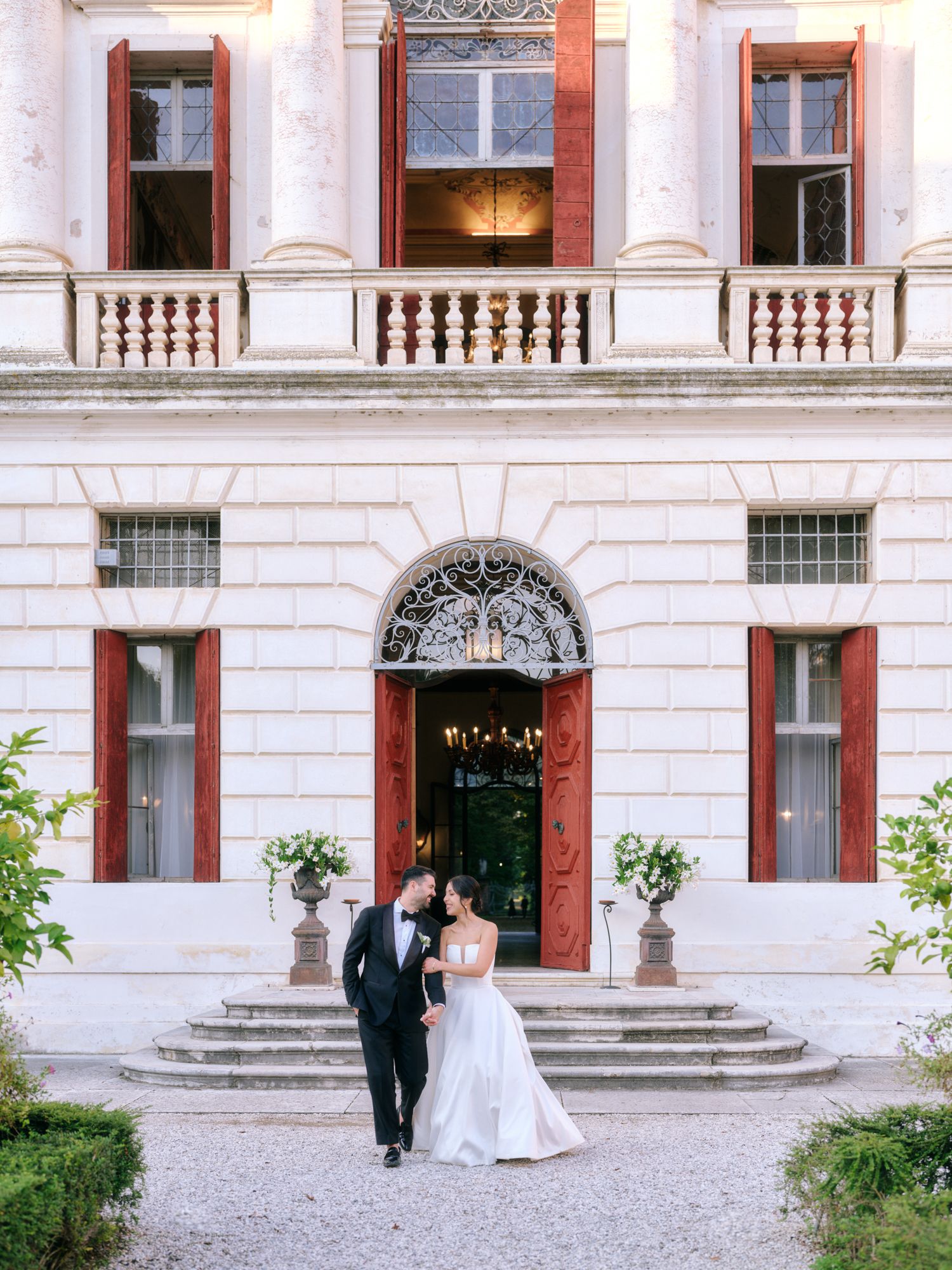A couple in formal attire walks along the elegant stone facade of a historic European-style building with grand steps.