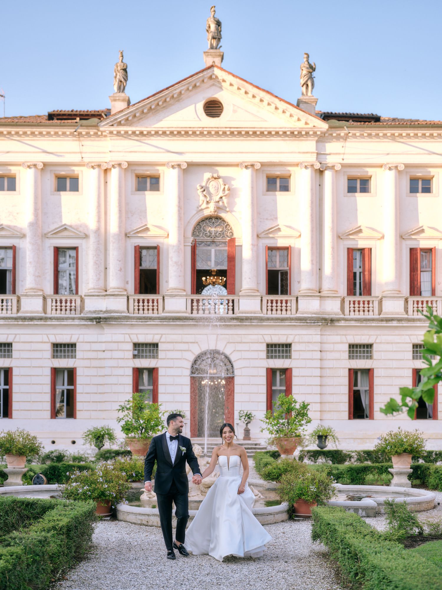 A couple strolls through a manicured garden courtyard of a luxurious white Italian villa with classical architecture.