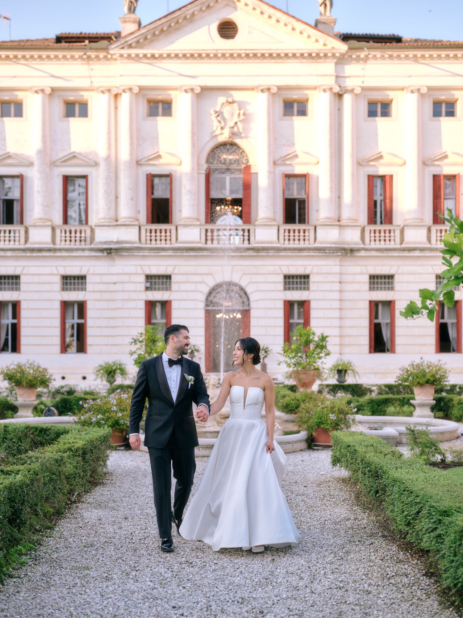 Elegant wedding couple strolls through manicured gardens of a grand historic palace with ornate architecture.