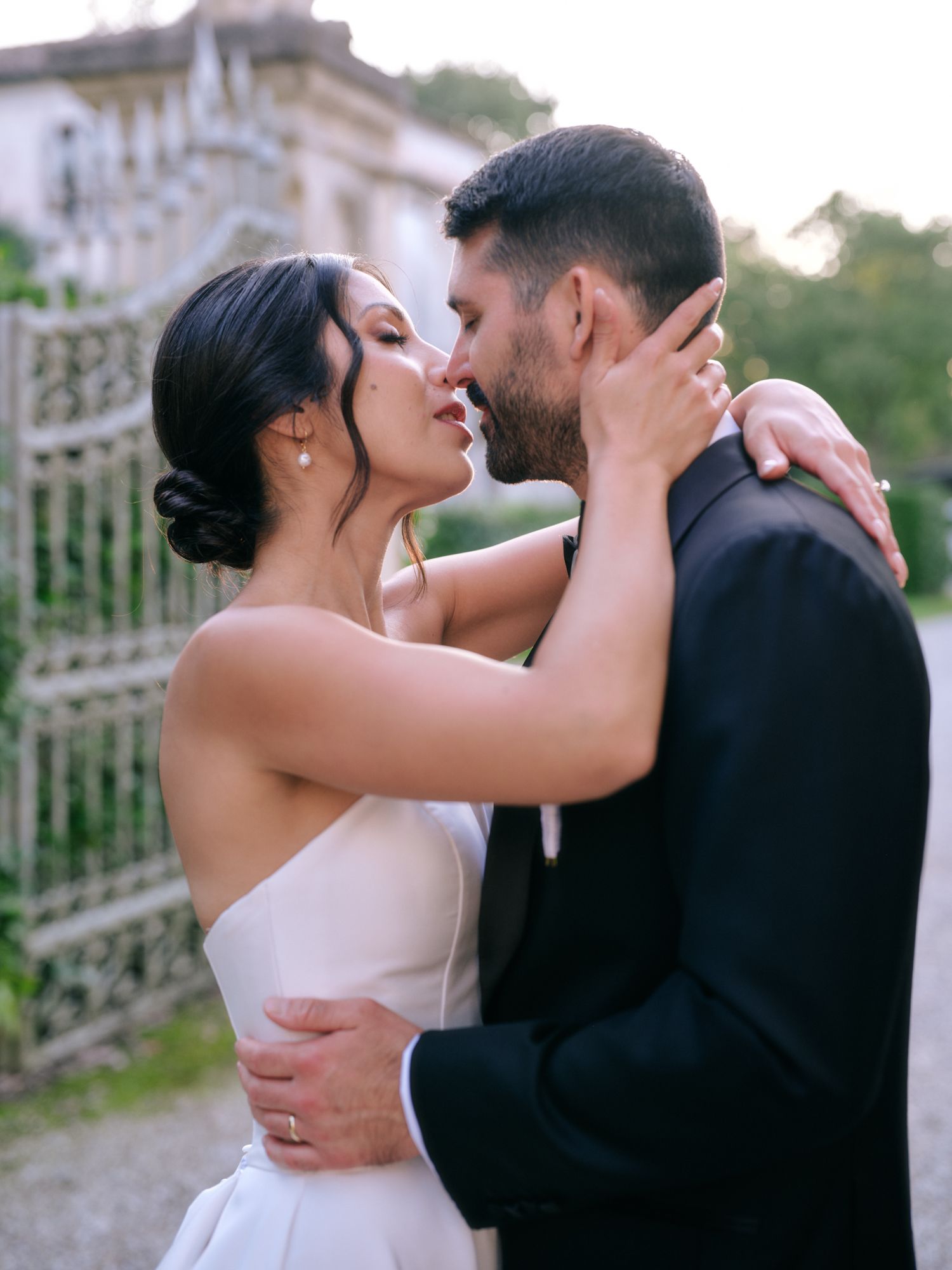 A romantic couple in wedding attire share an intimate kiss in front of an ornate garden gate.