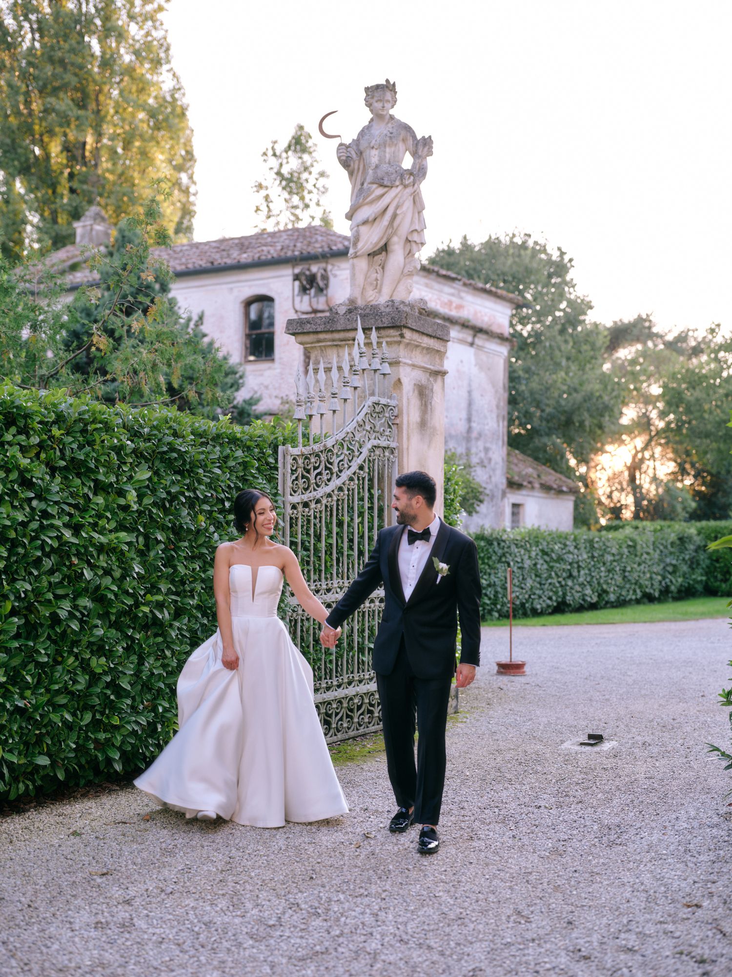 Elegant wedding couple shares romantic moments on a gravel path beside a majestic Italian villa at sunset.