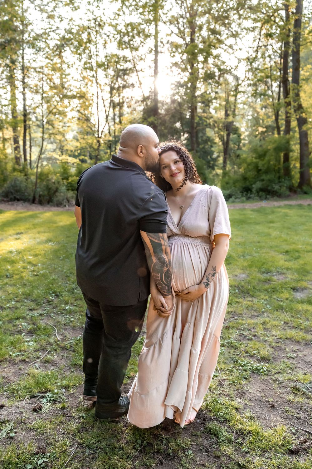 A romantic maternity photoshoot in a sunlit forest setting with a couple embracing in casual attire.