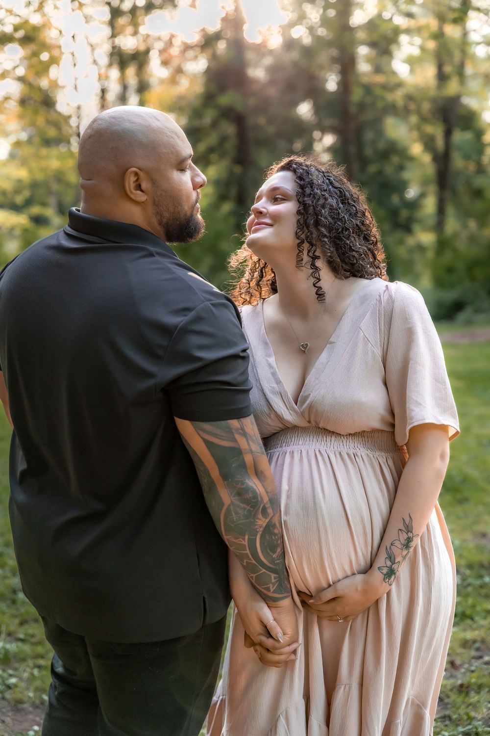 A couple shares an intimate moment together in a sunlit outdoor setting, displaying visible tattoos and affection.