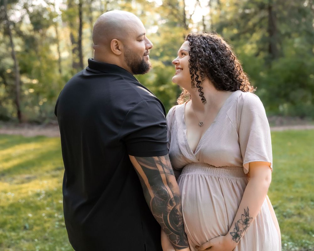 A couple in romantic embrace stands in a sunlit forest clearing wearing cream and black outfits.