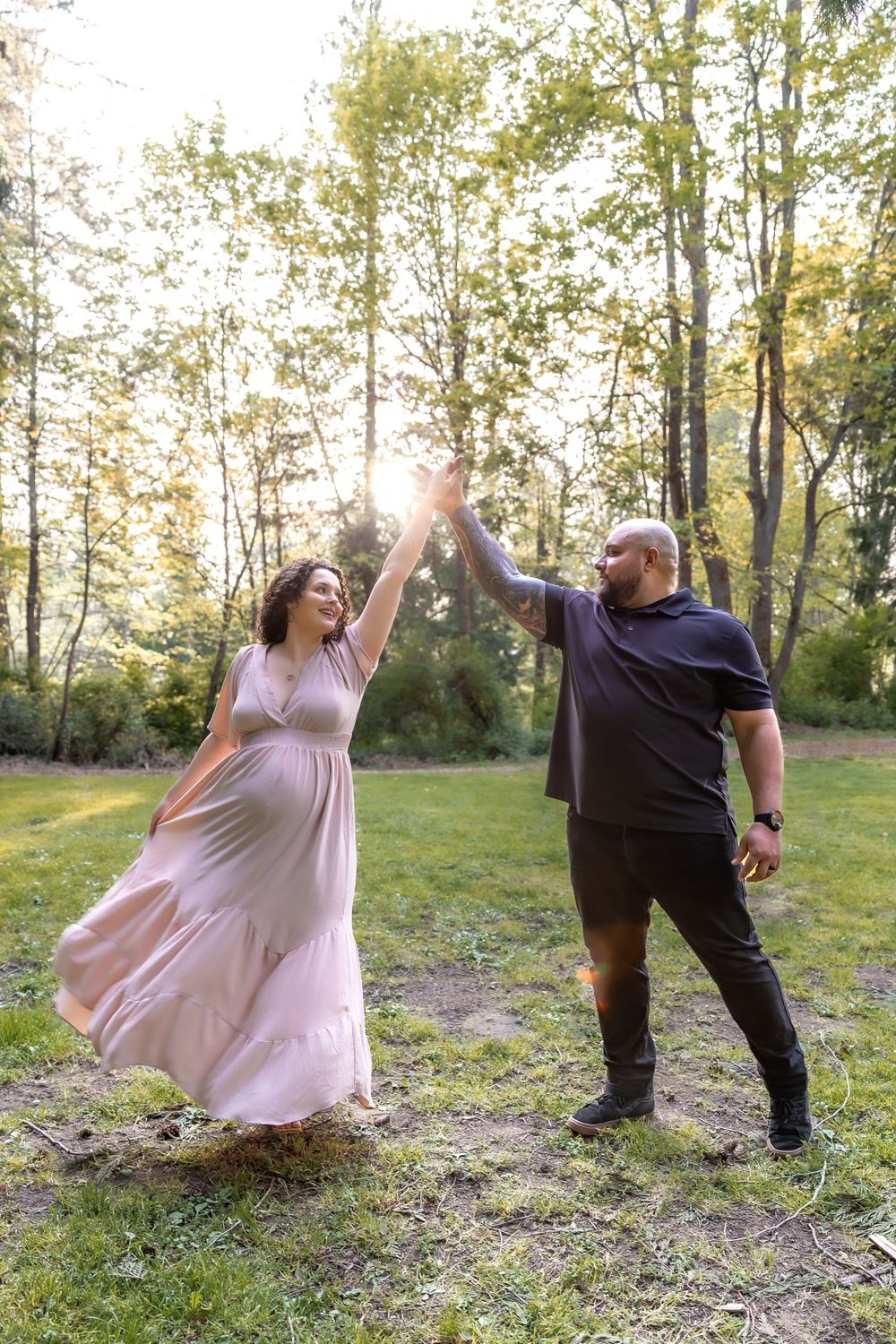 A couple dances playfully together in a sunlit meadow surrounded by trees on a warm afternoon.