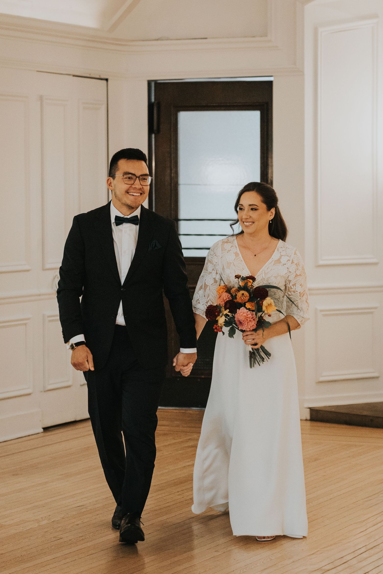 A joyful couple in formal wedding attire walks together across a bright hardwood floor in an elegant white room.