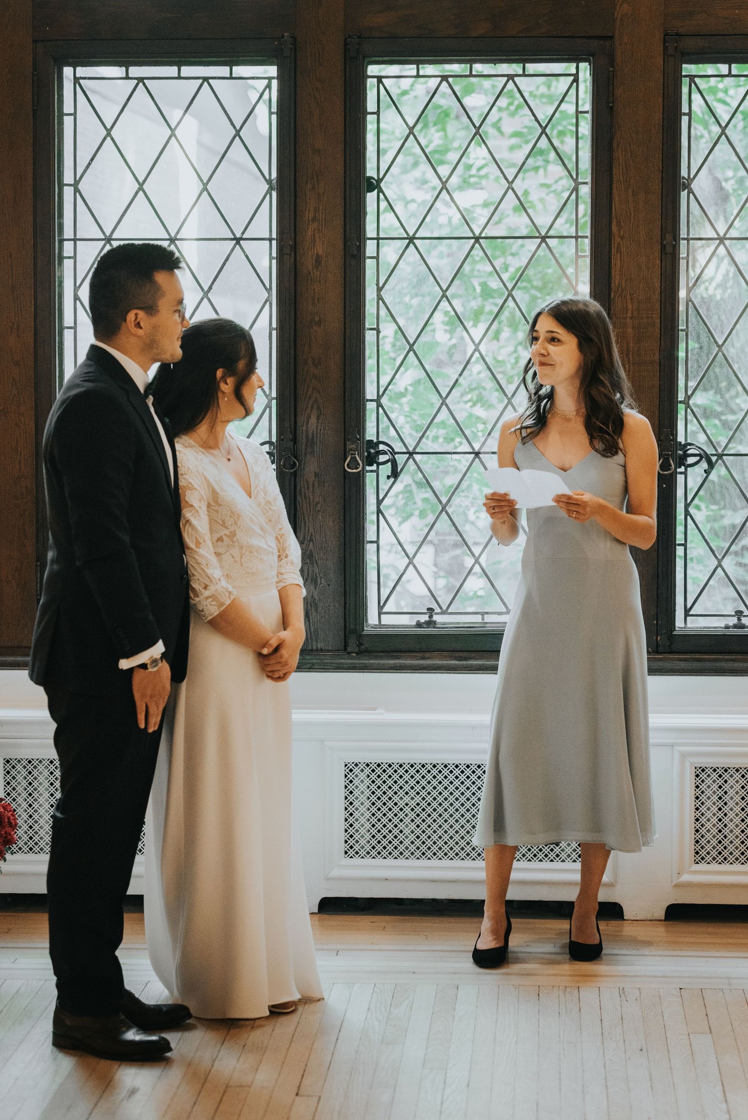 Wedding party members in black suits and light dresses stand together in an elegant room with diamond-paned windows.