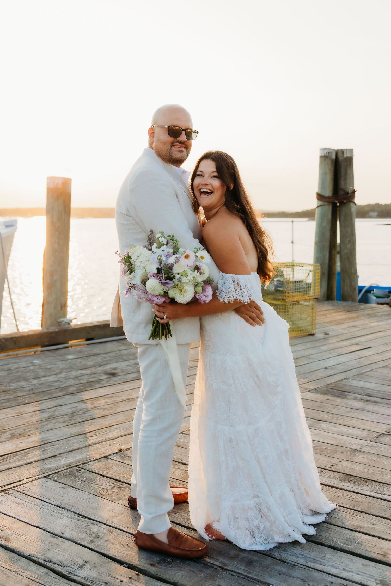 Happy couple shares intimate moments on a wooden pier at sunset while holding white floral bouquet.