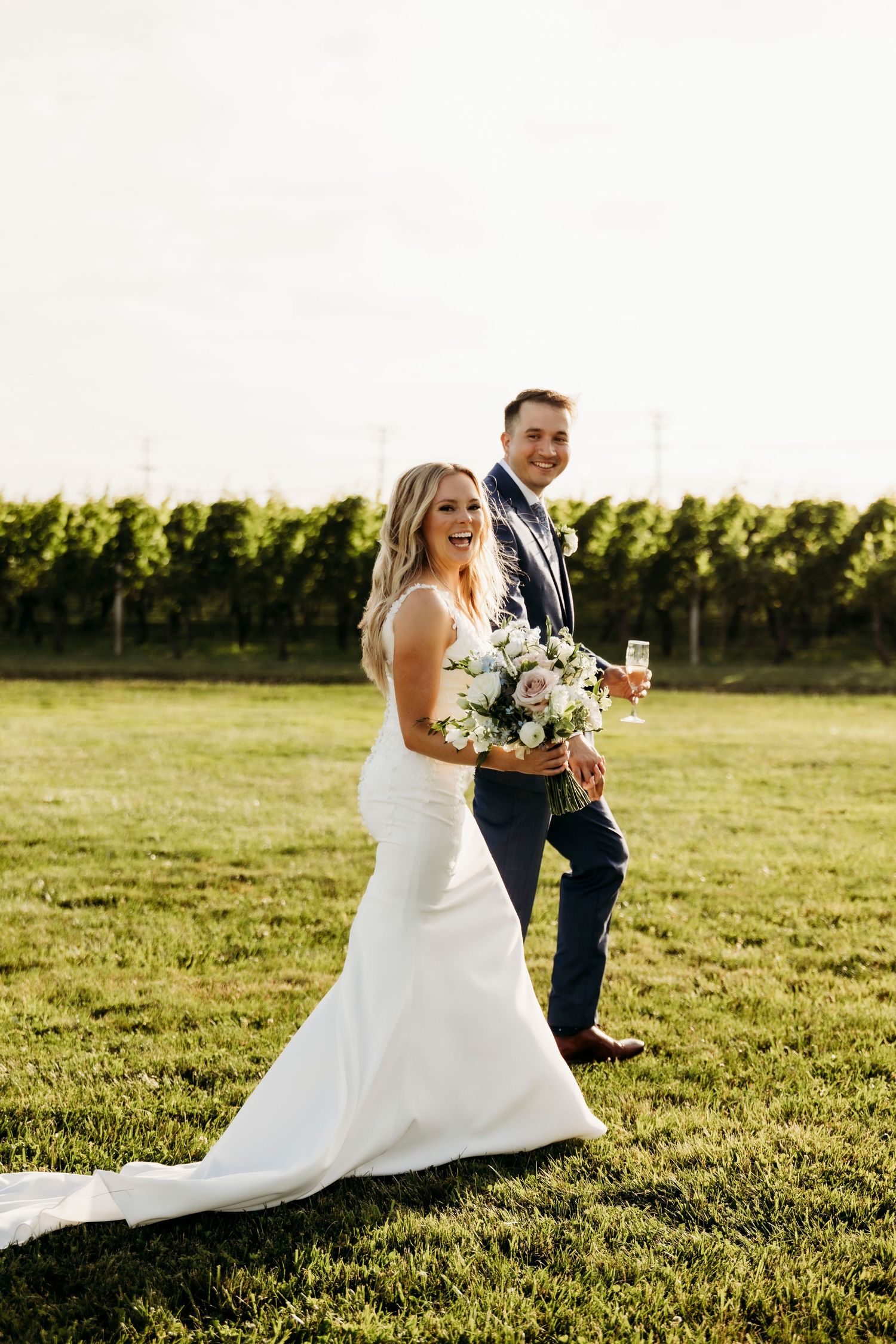 A couple in wedding attire sits together on a grassy field during golden hour sunset.