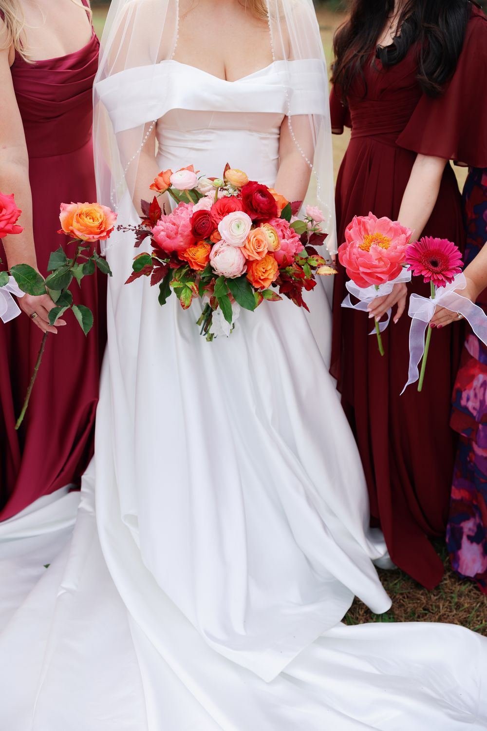 Bride in white wedding gown stands with bridesmaids in burgundy dresses holding vibrant coral and red floral bouquets.