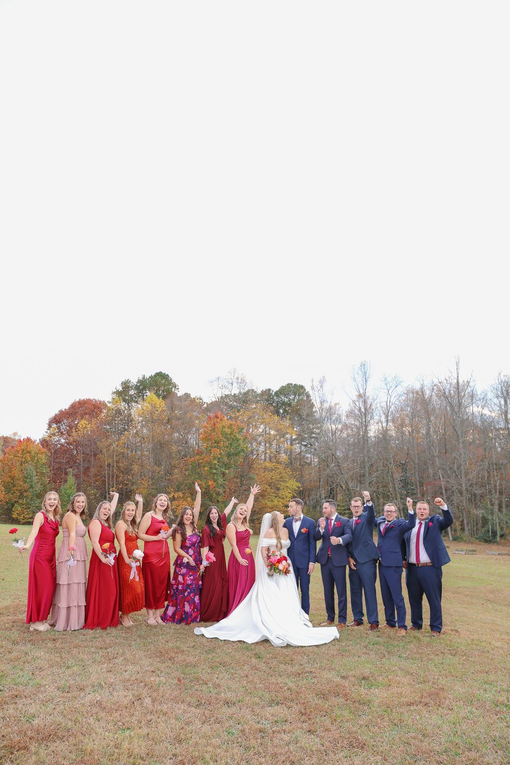 Wedding party in red dresses and navy suits poses together on a grassy hill during autumn.