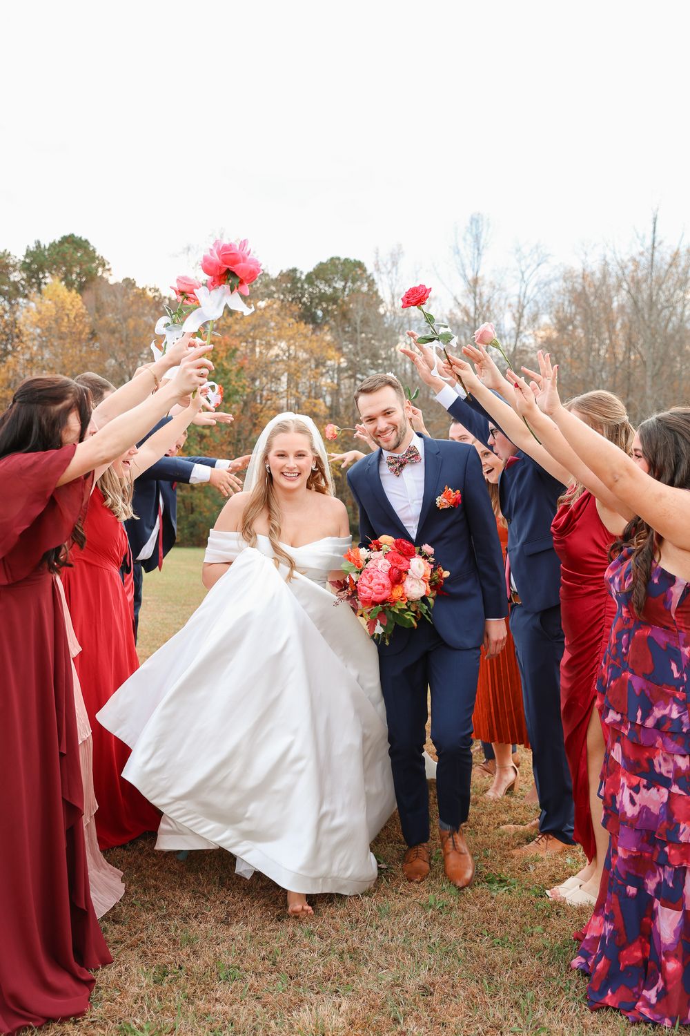 Wedding party celebrates with flower petals as newlyweds walk under raised arms on autumn day outdoors.