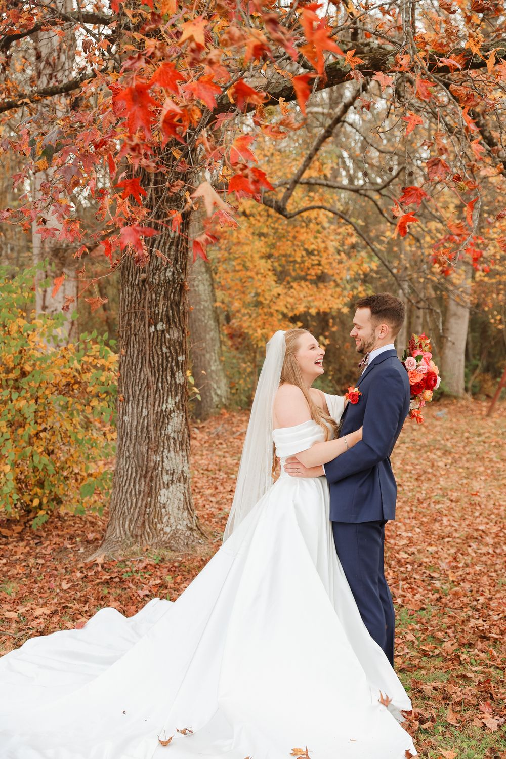 A romantic autumn wedding photo with a couple embracing under vibrant red maple leaves in a wooded setting.