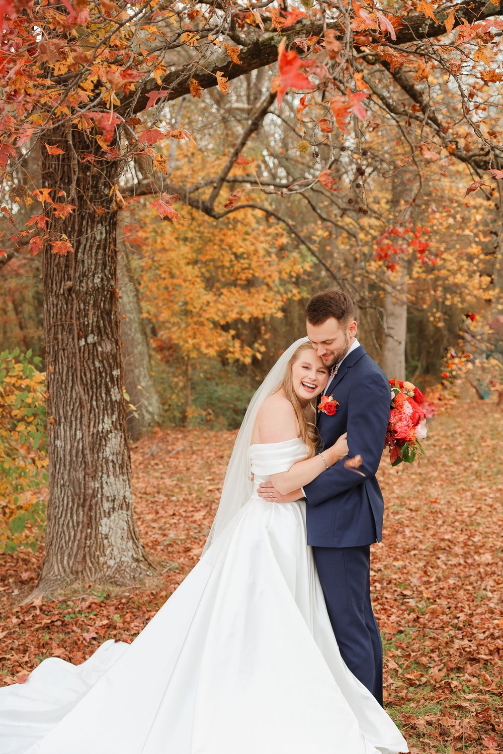 A couple shares an intimate moment surrounded by vibrant fall foliage as her white wedding dress trails on autumn leaves.